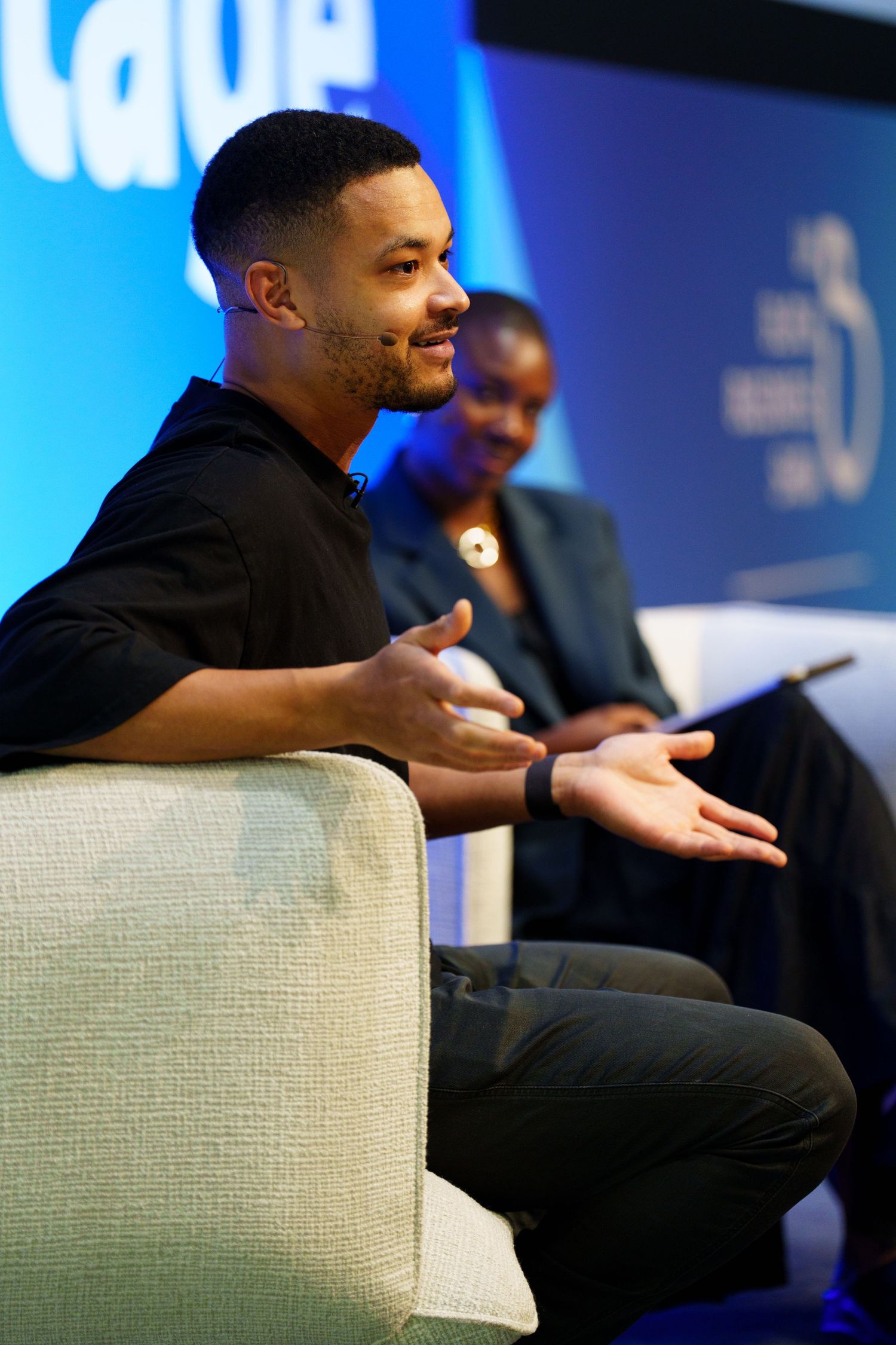 A speaker sits in a light colored armchair during a panel discussion with blue stage lighting in the background.