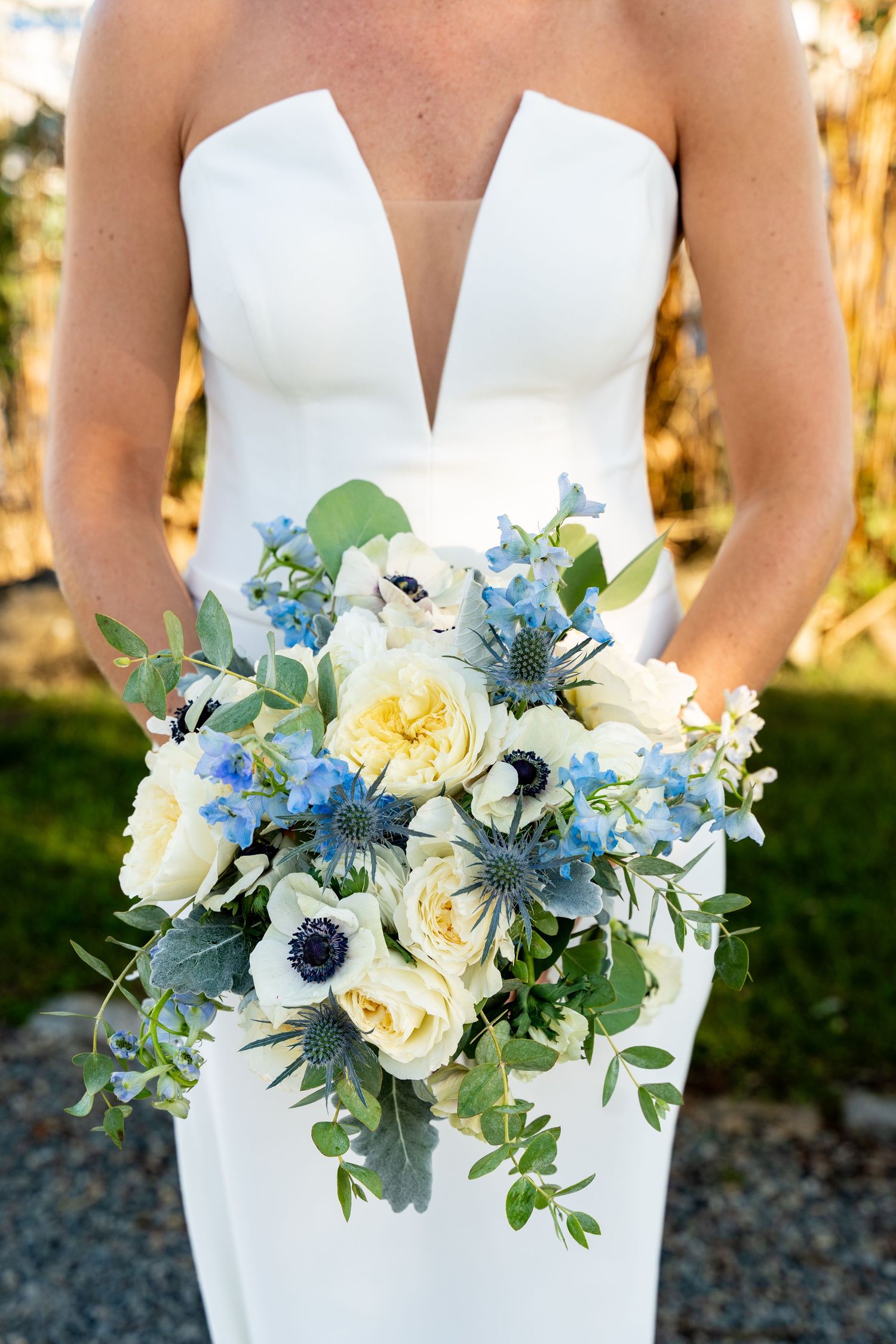 An elegant bridal bouquet features blue and white flowers with green eucalyptus against a white wedding dress.