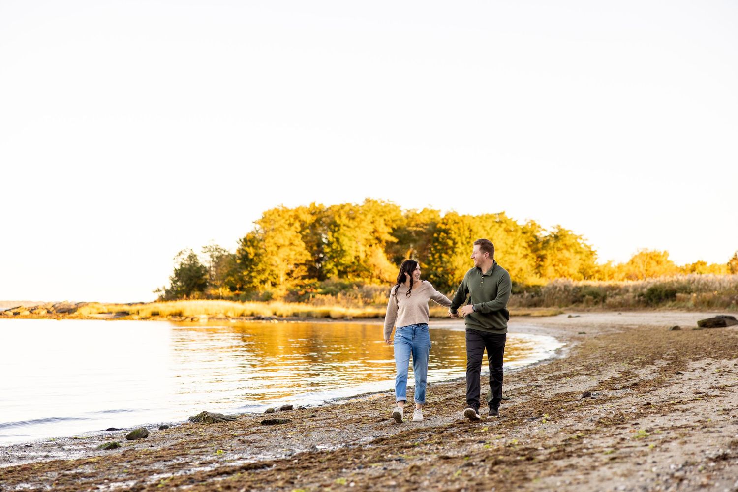 A couple walks along a beach shoreline with golden fall foliage in the background.