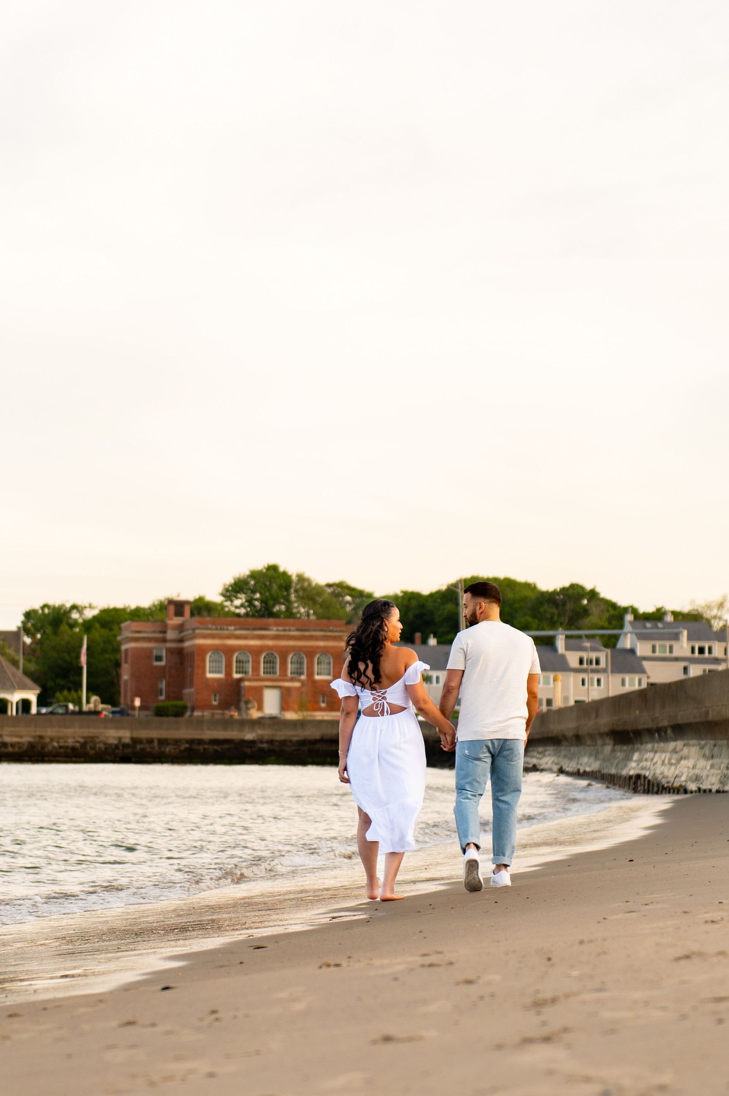A couple walks along the beach with buildings visible in the background at sunset.