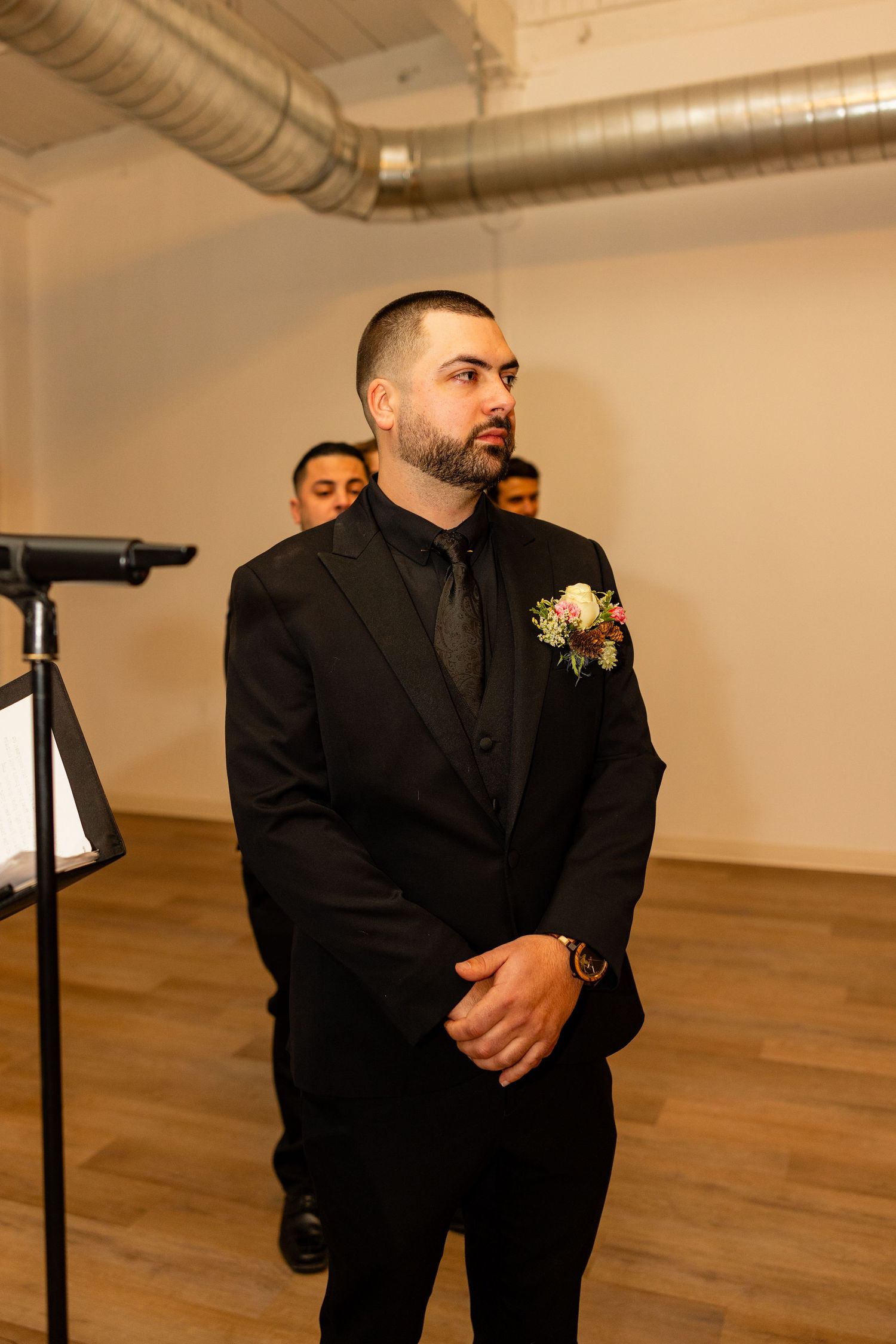 A groom in a black suit stands attentively during the wedding ceremony.