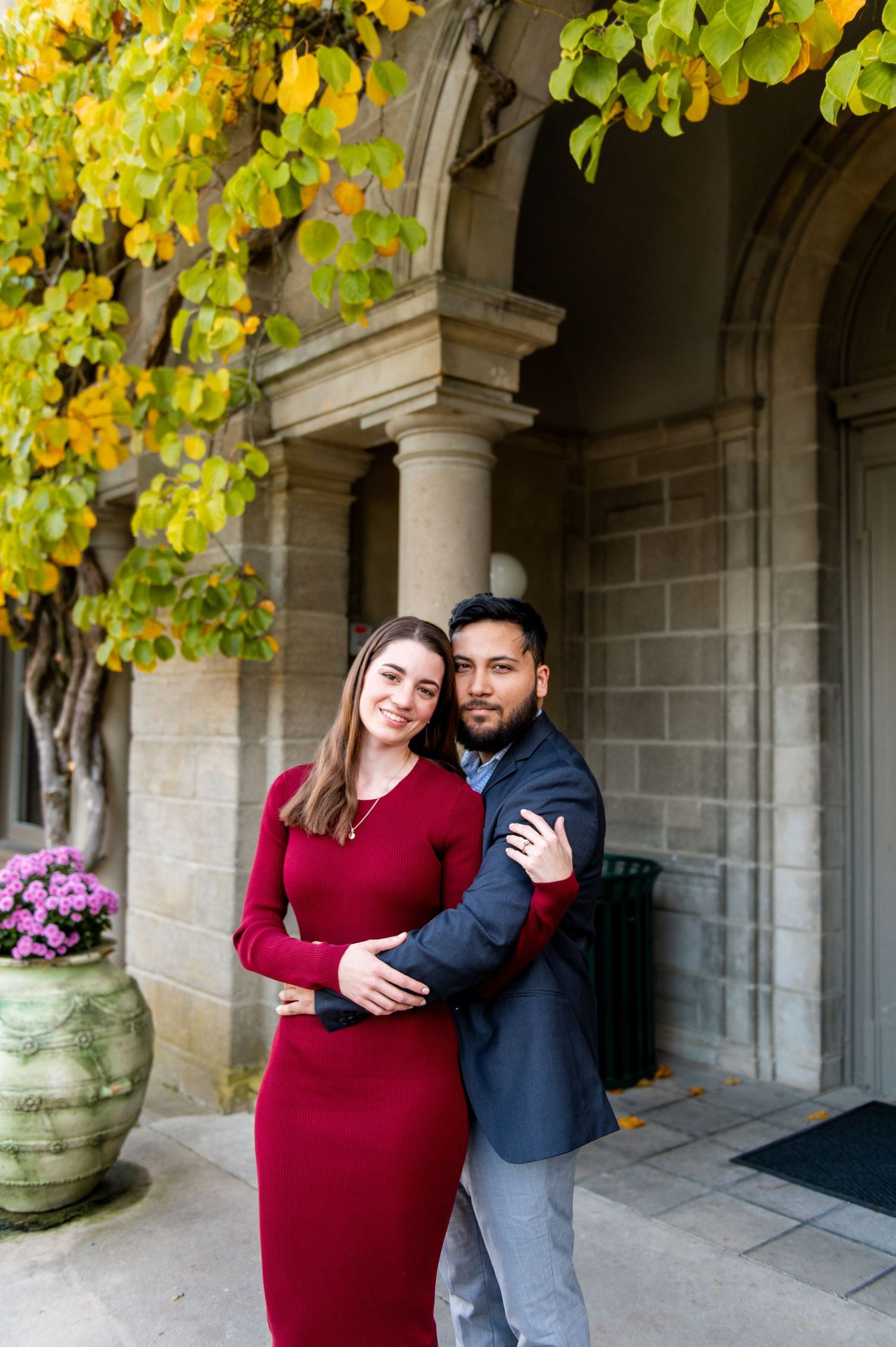 A couple embraces in front of a stone building with autumn foliage and decorative columns.