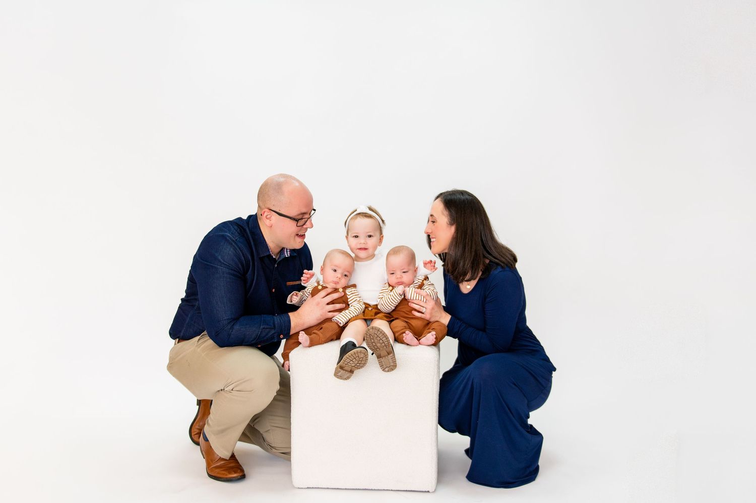 Family wearing coordinated navy and khaki outfits pose together with small children on white box.