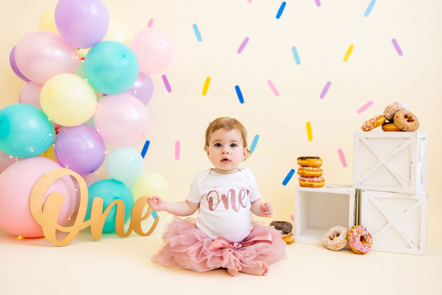 First birthday setup with baby in tutu, pastel balloons, and gold 'one' sign.