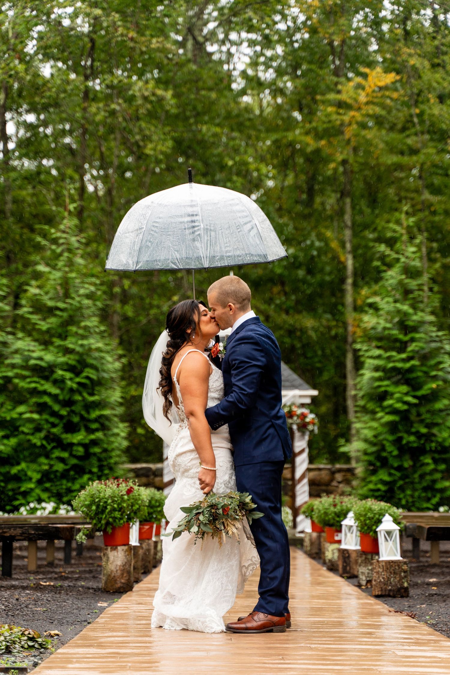 Couple kisses on rustic wooden walkway under clear umbrella with lantern decorations.