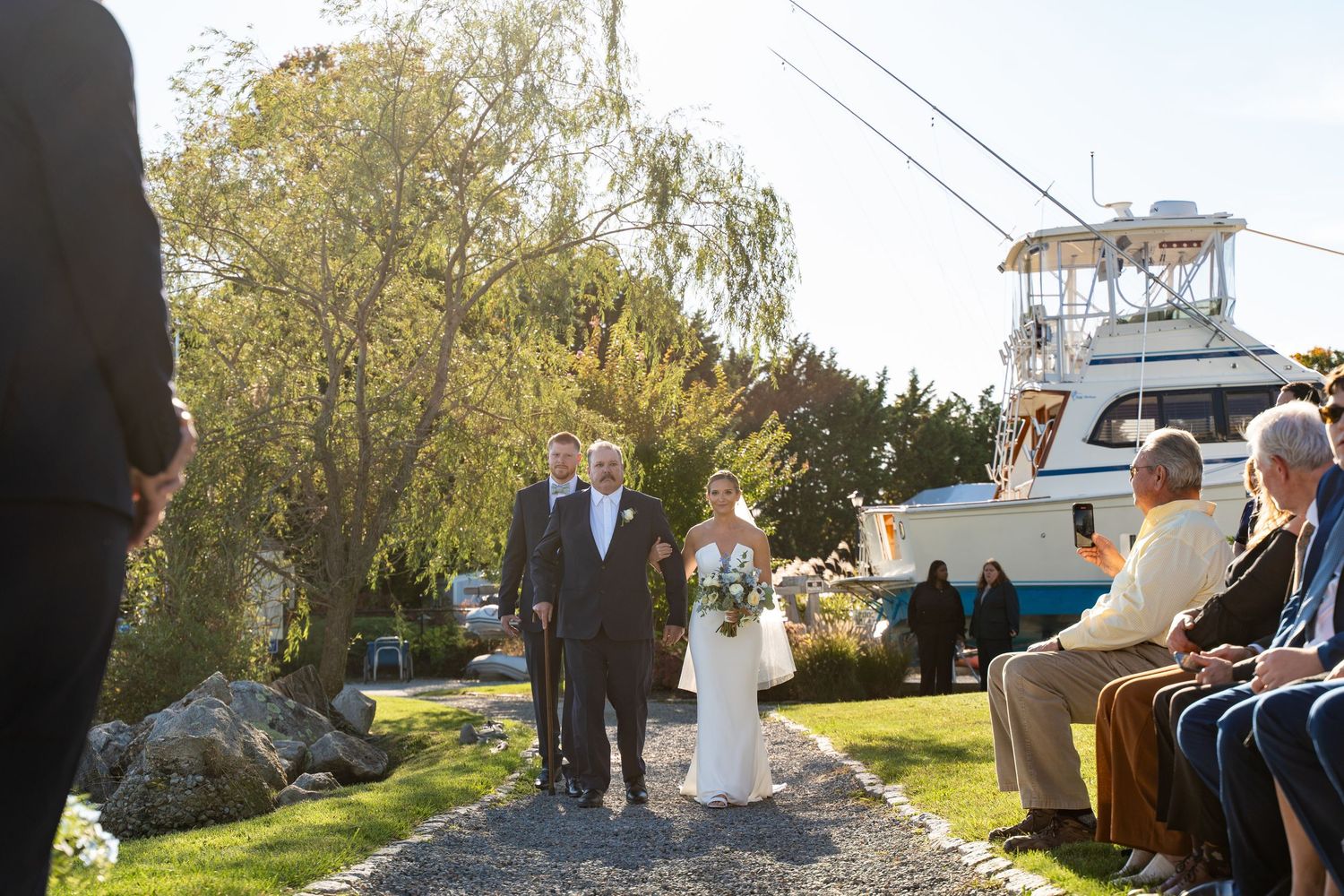 Wedding processional on scenic coastal path with boat in background.
