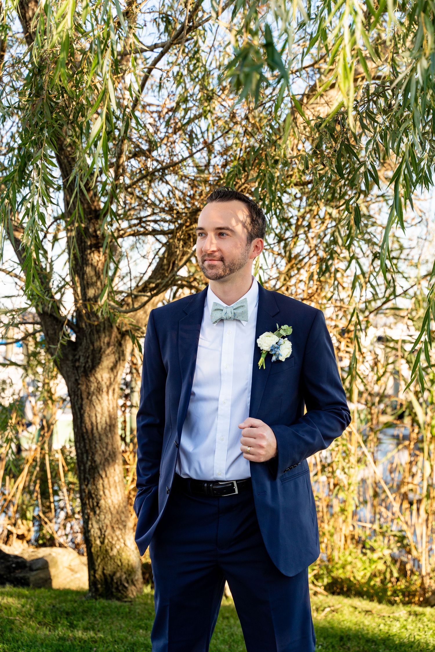 Groom in navy suit and bow tie poses under willow tree branches.