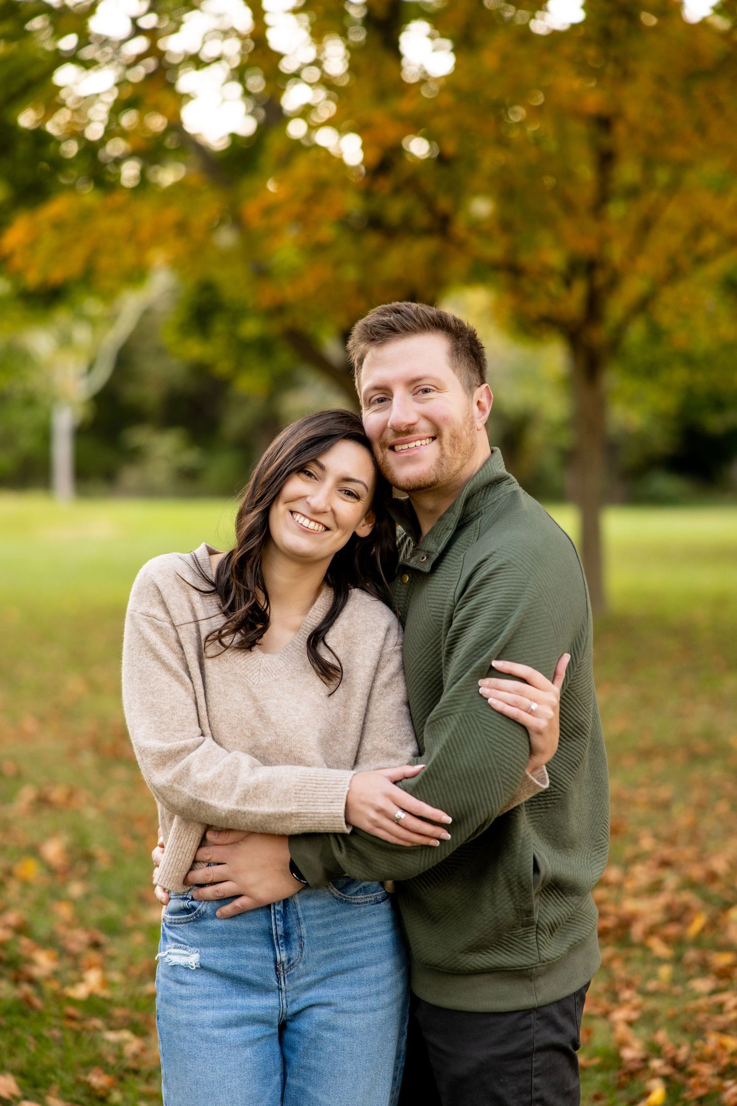 A couple embraces while smiling at the camera in a park with autumn trees in the background.