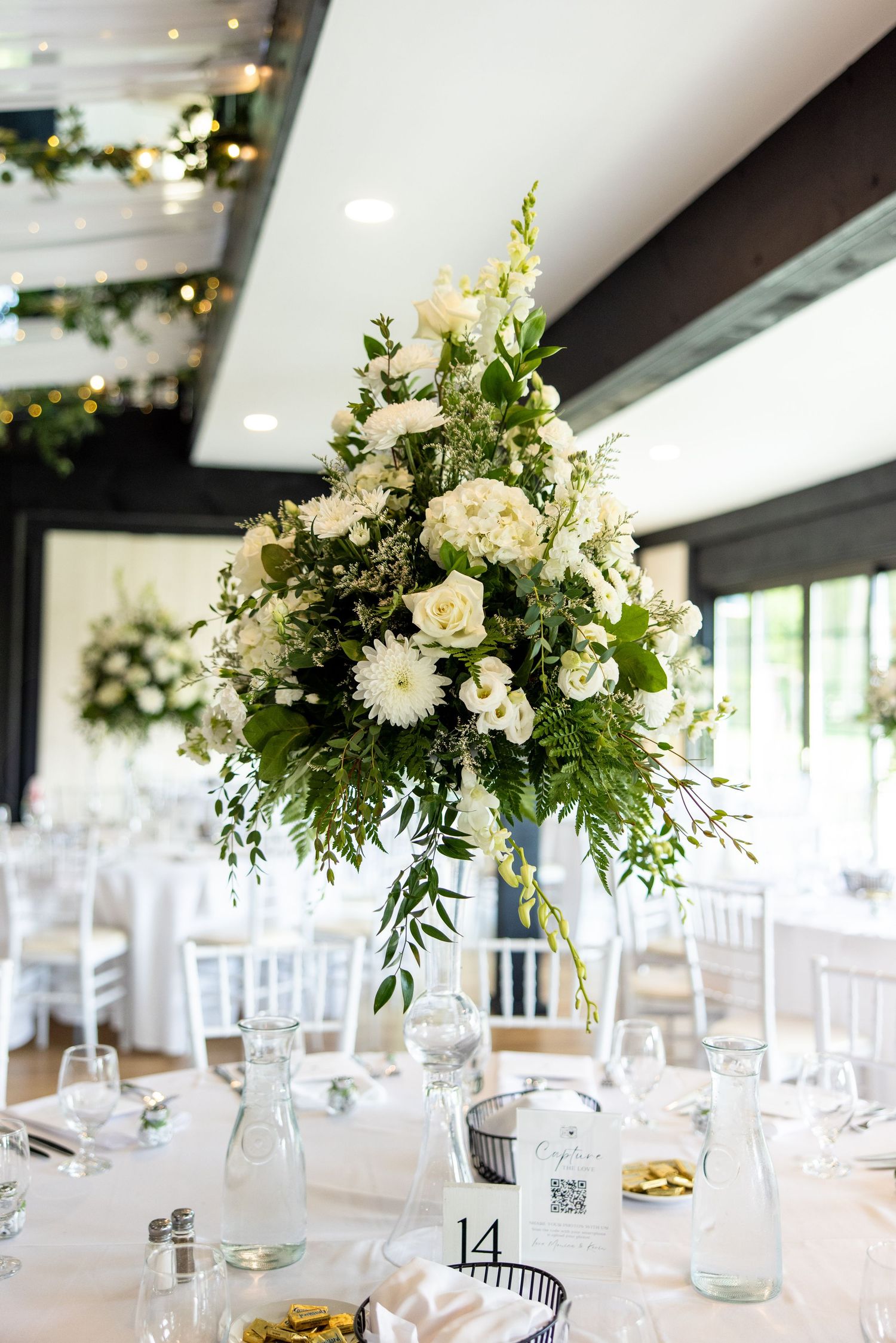 Elegant white floral centerpiece with roses and greenery in tall vase on round reception table with place settings.