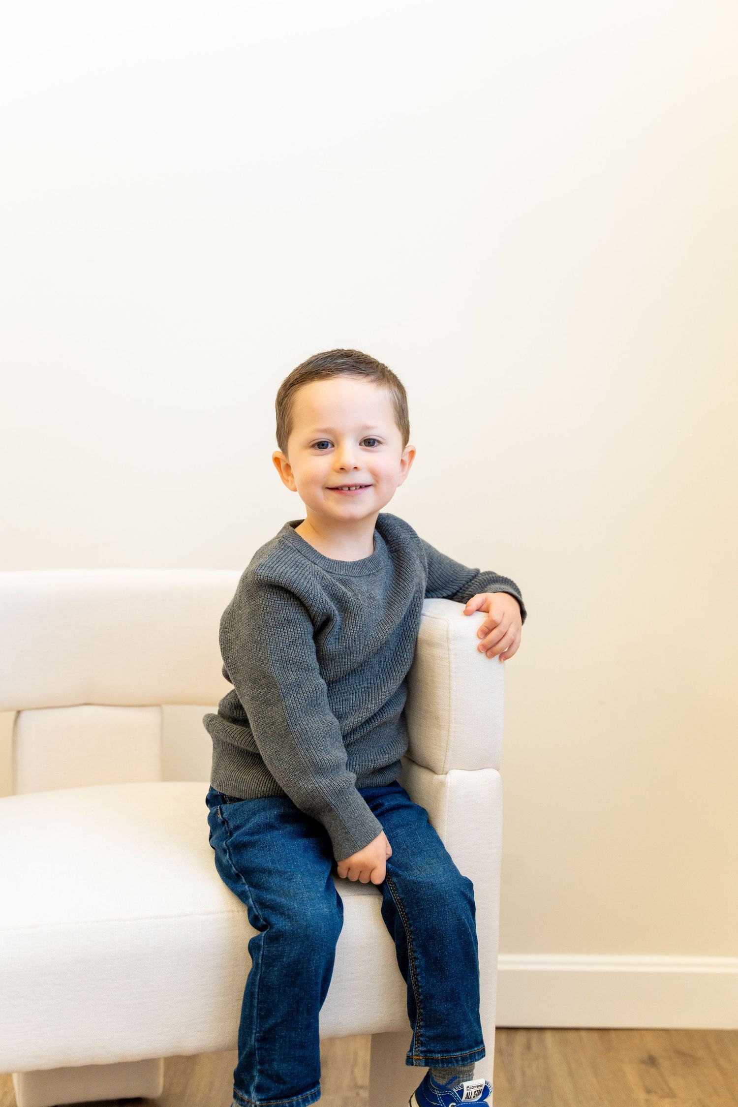 Child wearing gray sweater and jeans sitting casually on white couch.
