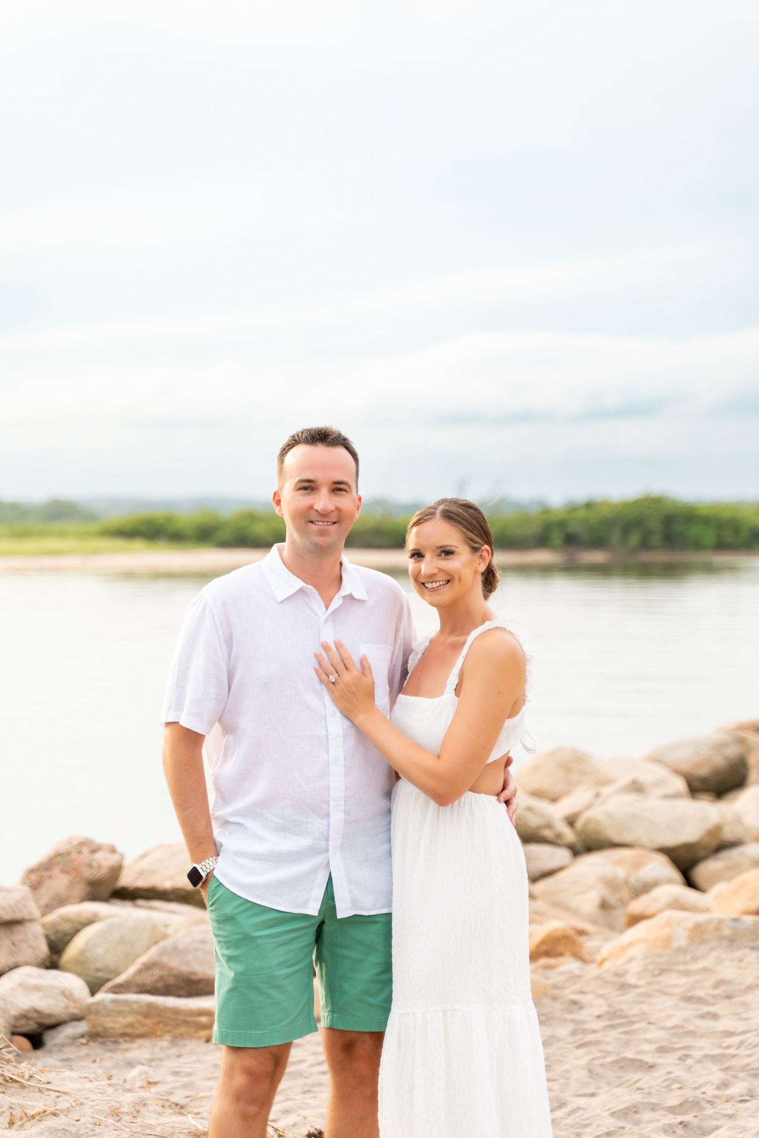 A couple stands together on a rocky beach shoreline during an engagement photoshoot.