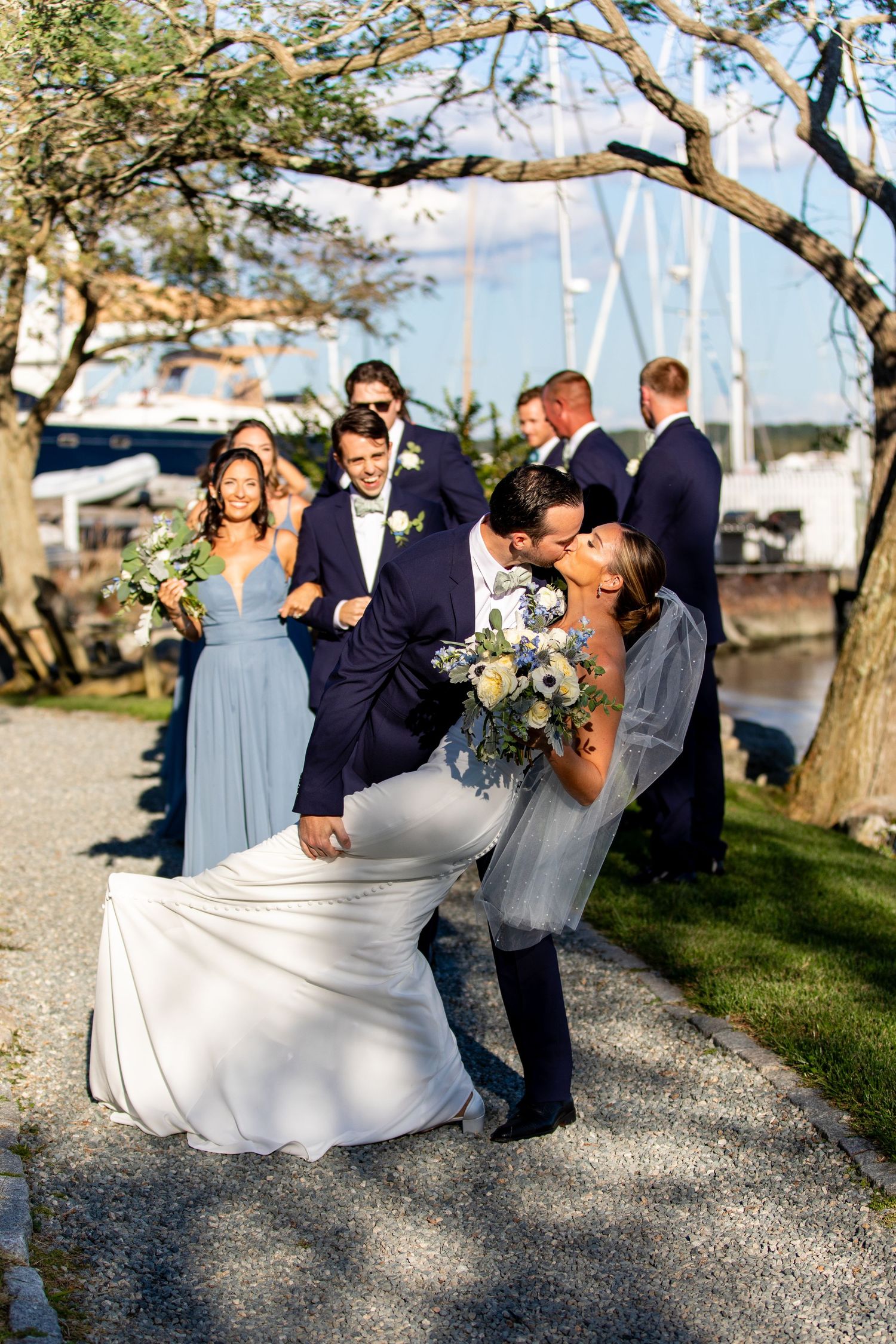 Wedding party in navy and light blue attire posing on gravel path by waterfront.