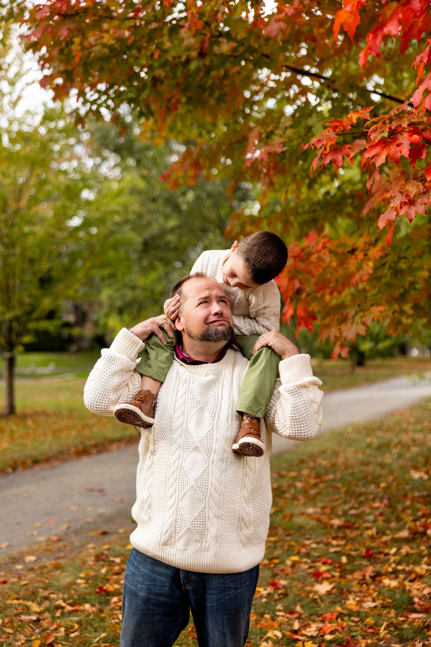 Child playfully rides on parent's shoulders under vibrant red maple leaves.