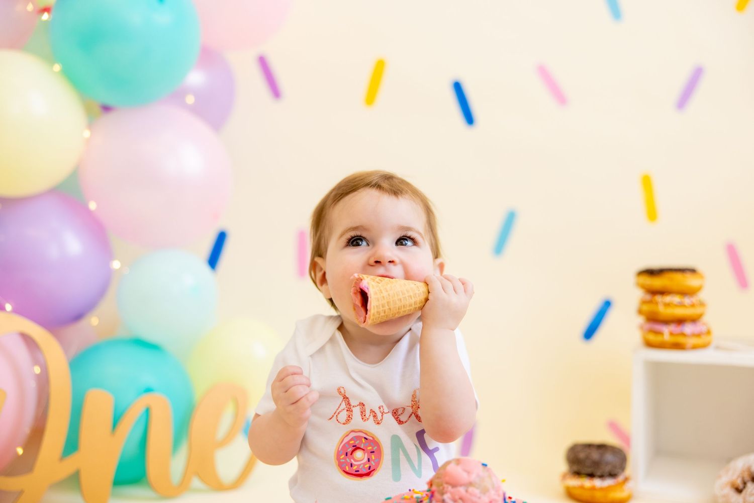 Baby enjoying cake at first birthday party with colorful confetti wall and balloon decorations.