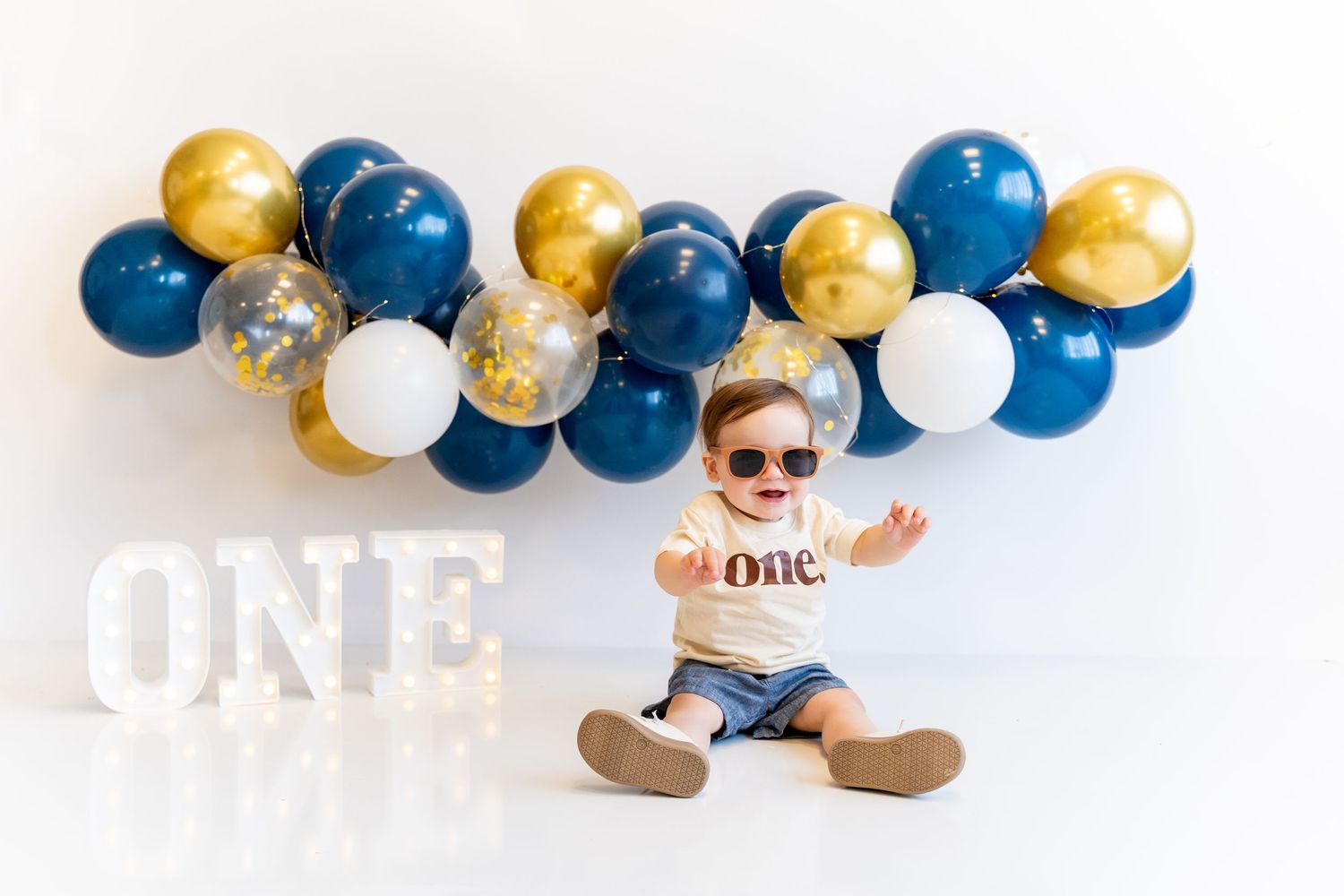 First birthday photo with baby wearing sunglasses under blue and gold balloon arch.