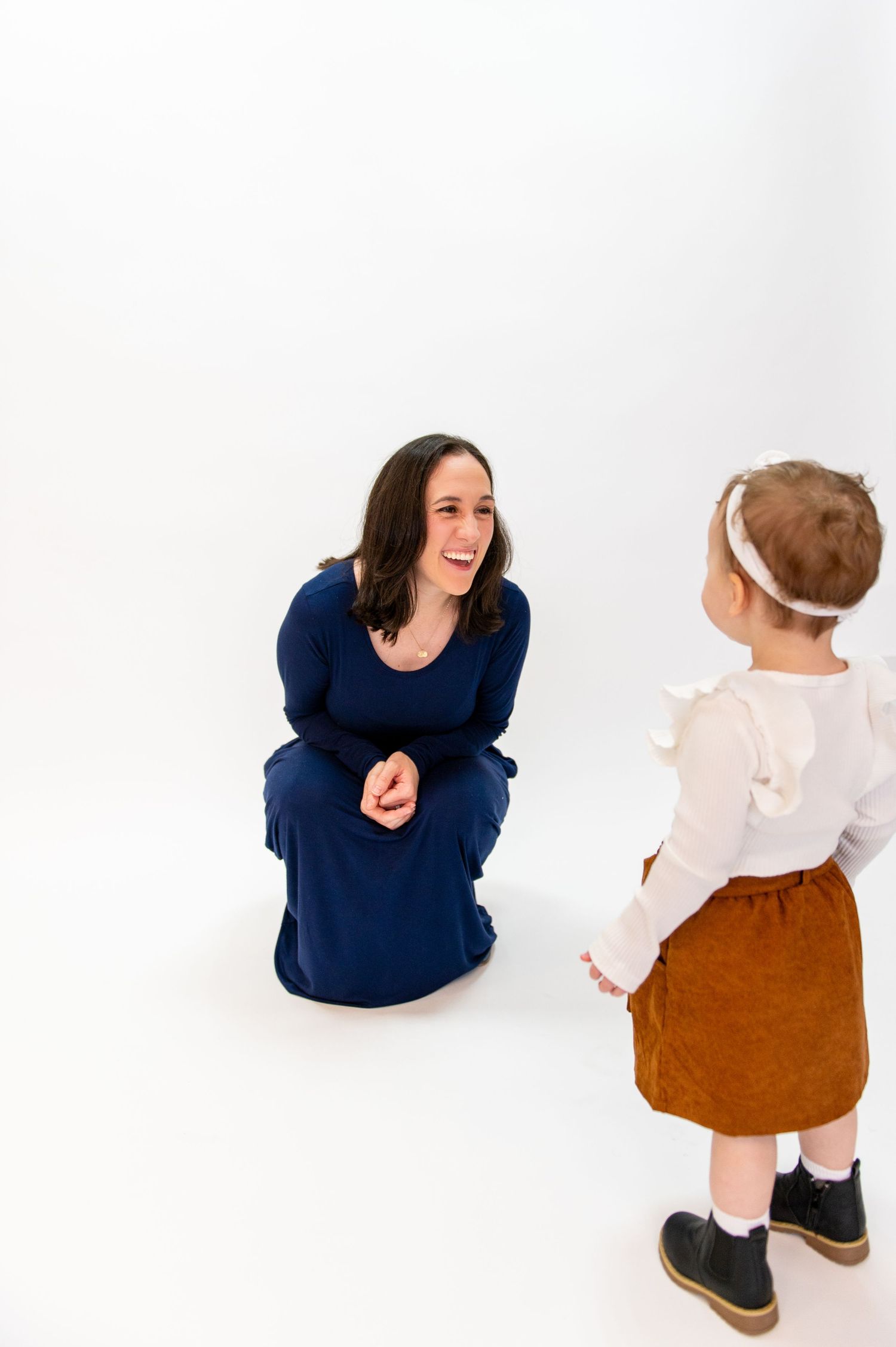 Woman in navy dress kneels to interact with child in white top and brown skirt.