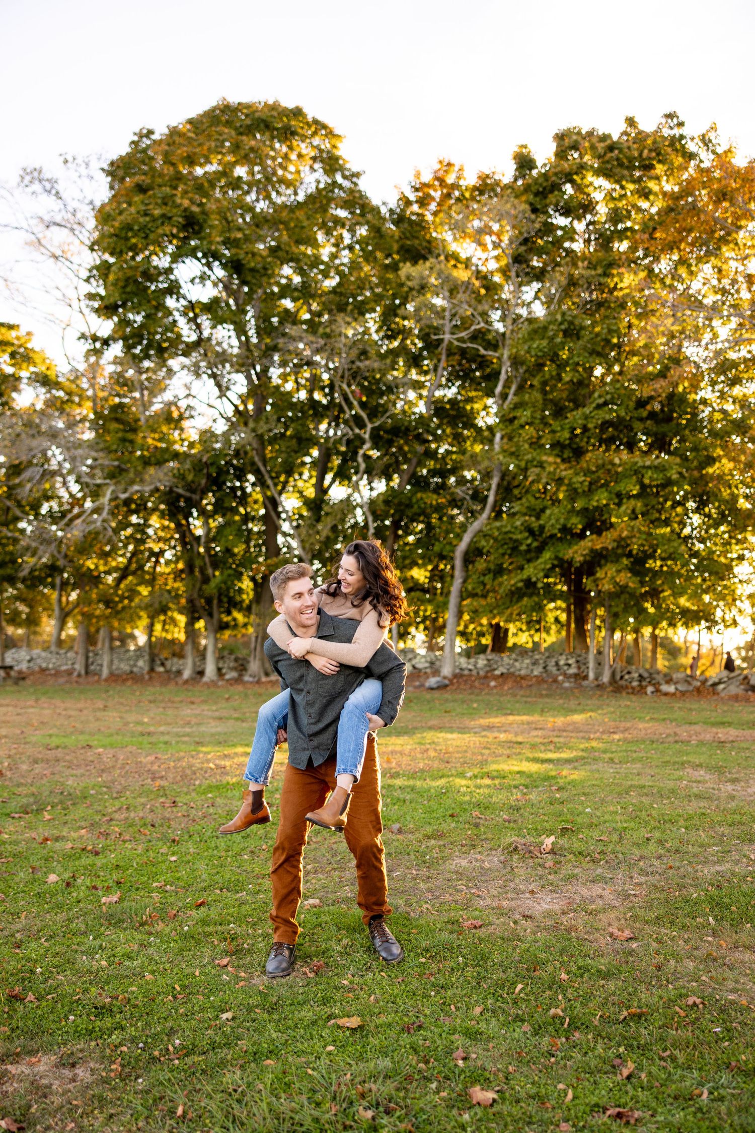 A playful couple shares a piggyback ride in a park surrounded by fall foliage.