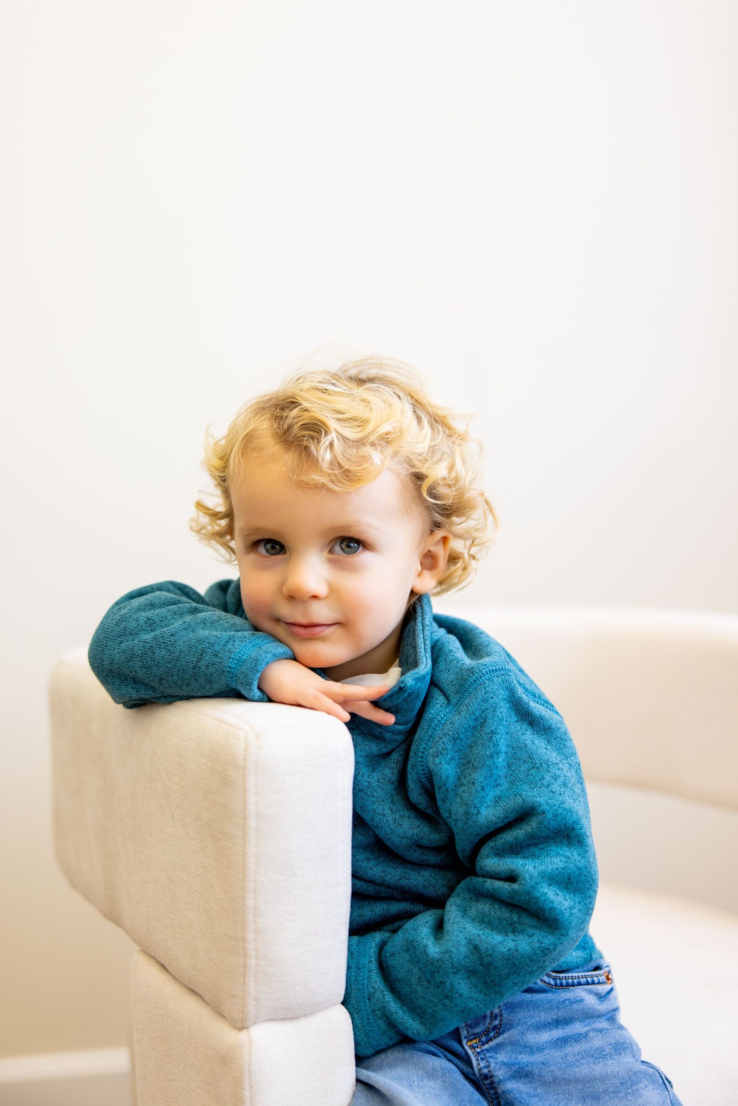 A toddler in teal sweater leans on white armchair cushion in minimalist setting.