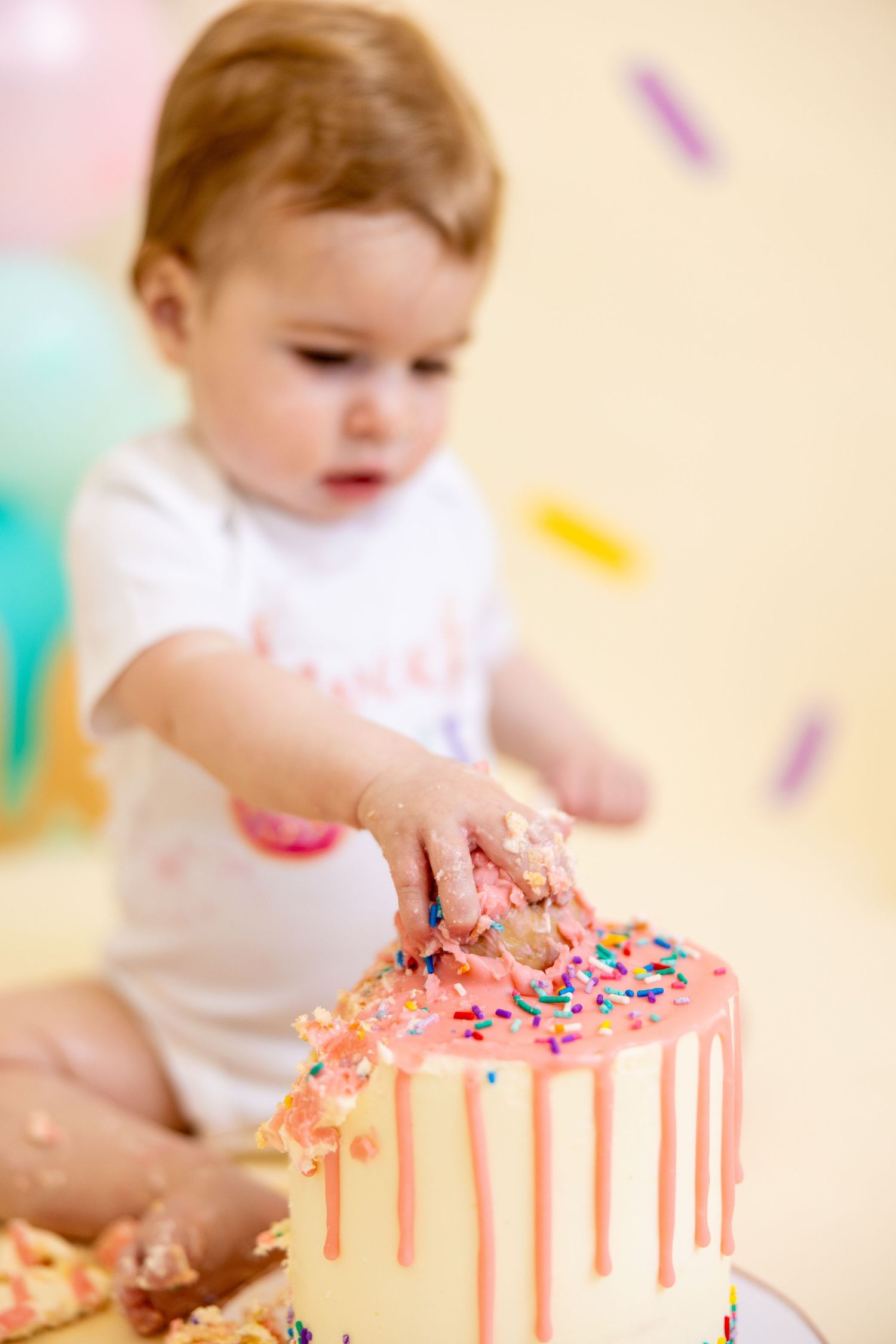 Baby reaches for pink drip cake decorated with colorful sprinkles during cake smash photoshoot.