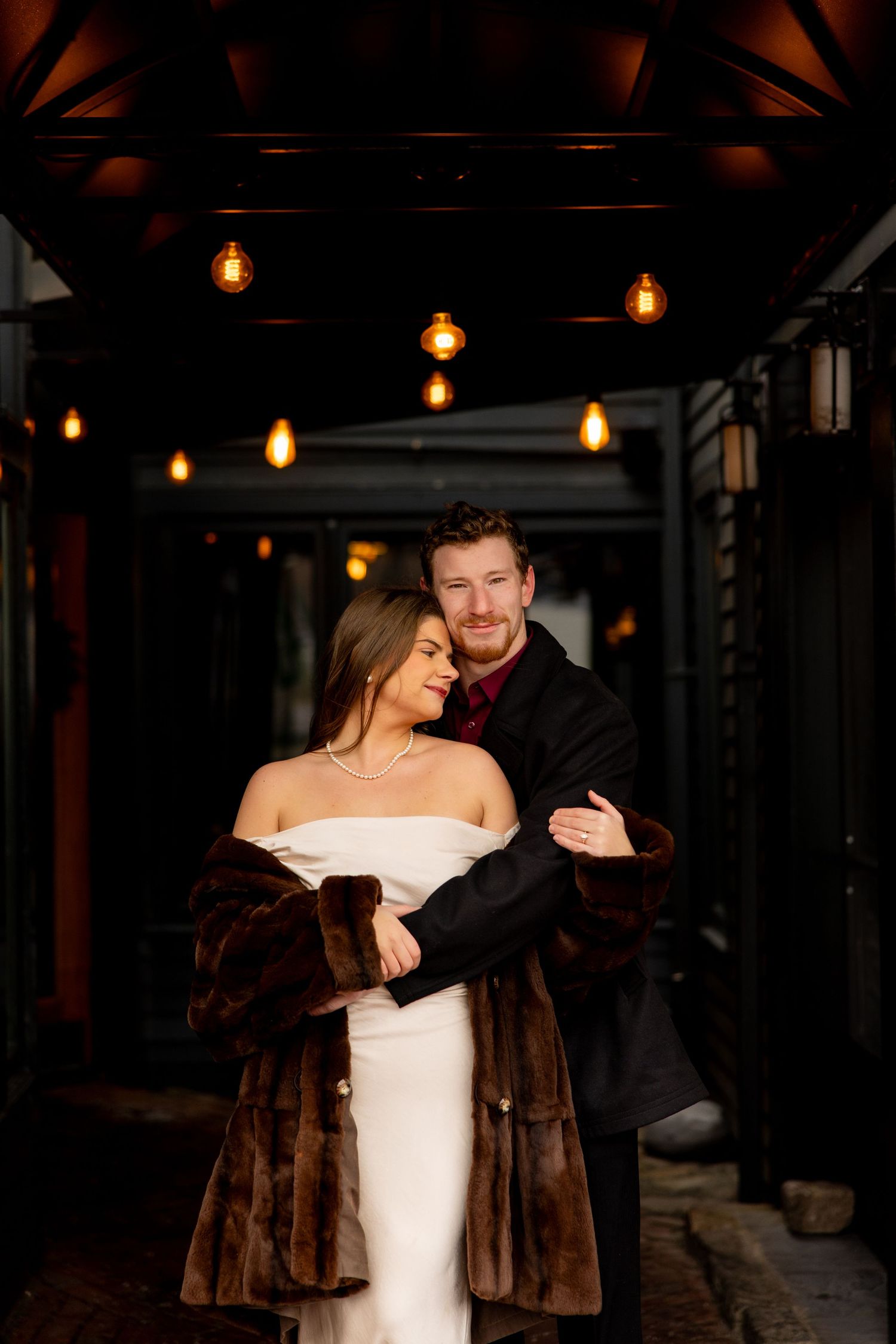 Romantic couple embracing under warm pendant lights in a dark hallway setting.