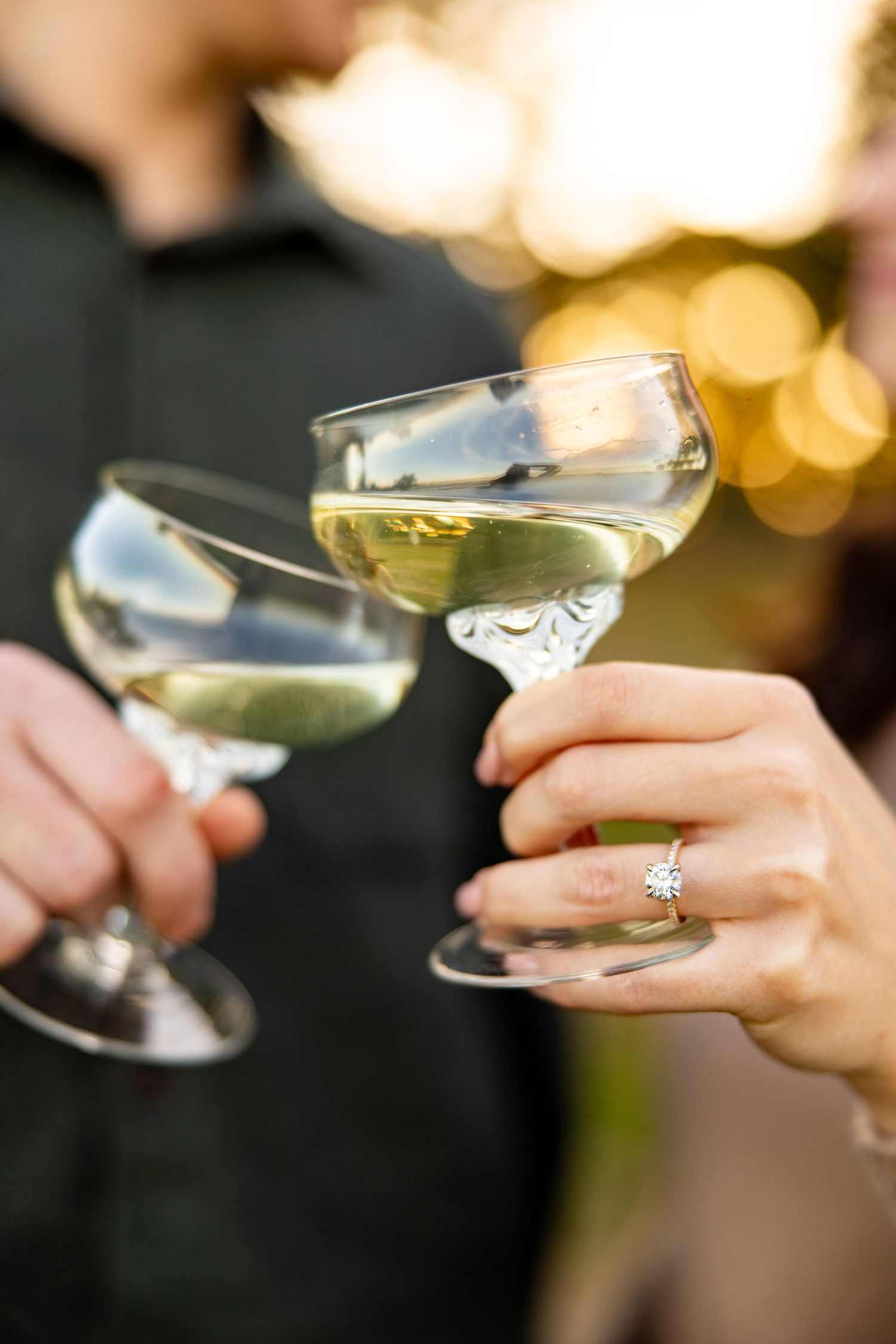 Two champagne glasses clink together with a sparkling engagement ring visible.