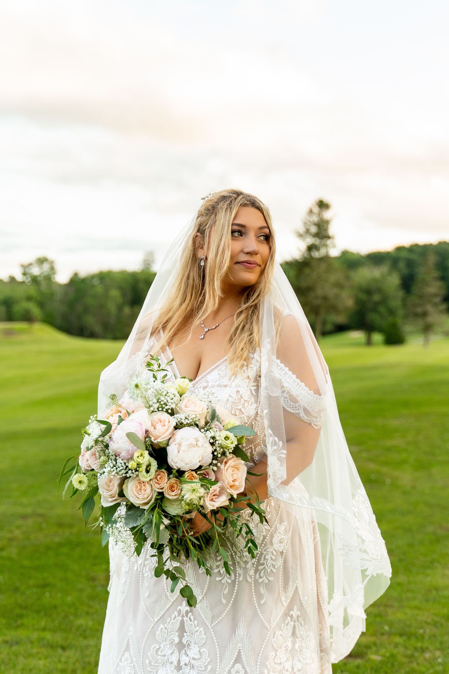 A bride in a lace wedding dress holds a blush and white bouquet on a green lawn.