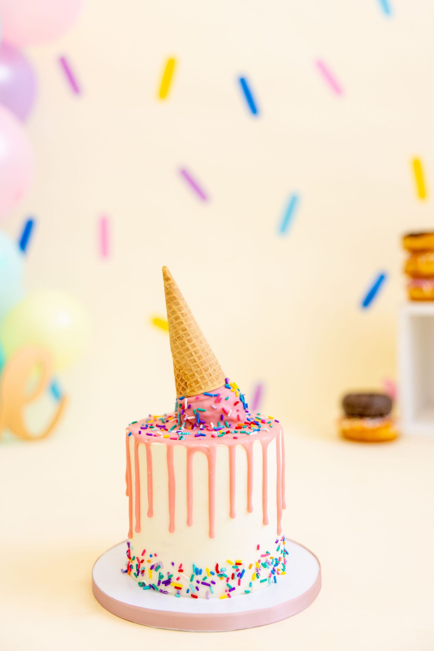 Drip cake decorated with sprinkles and ice cream cone topper against pastel party backdrop.
