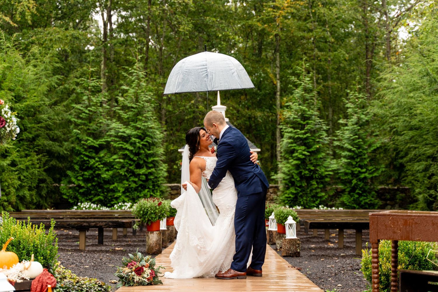 Rainy garden wedding ceremony features couple embracing under transparent umbrella.