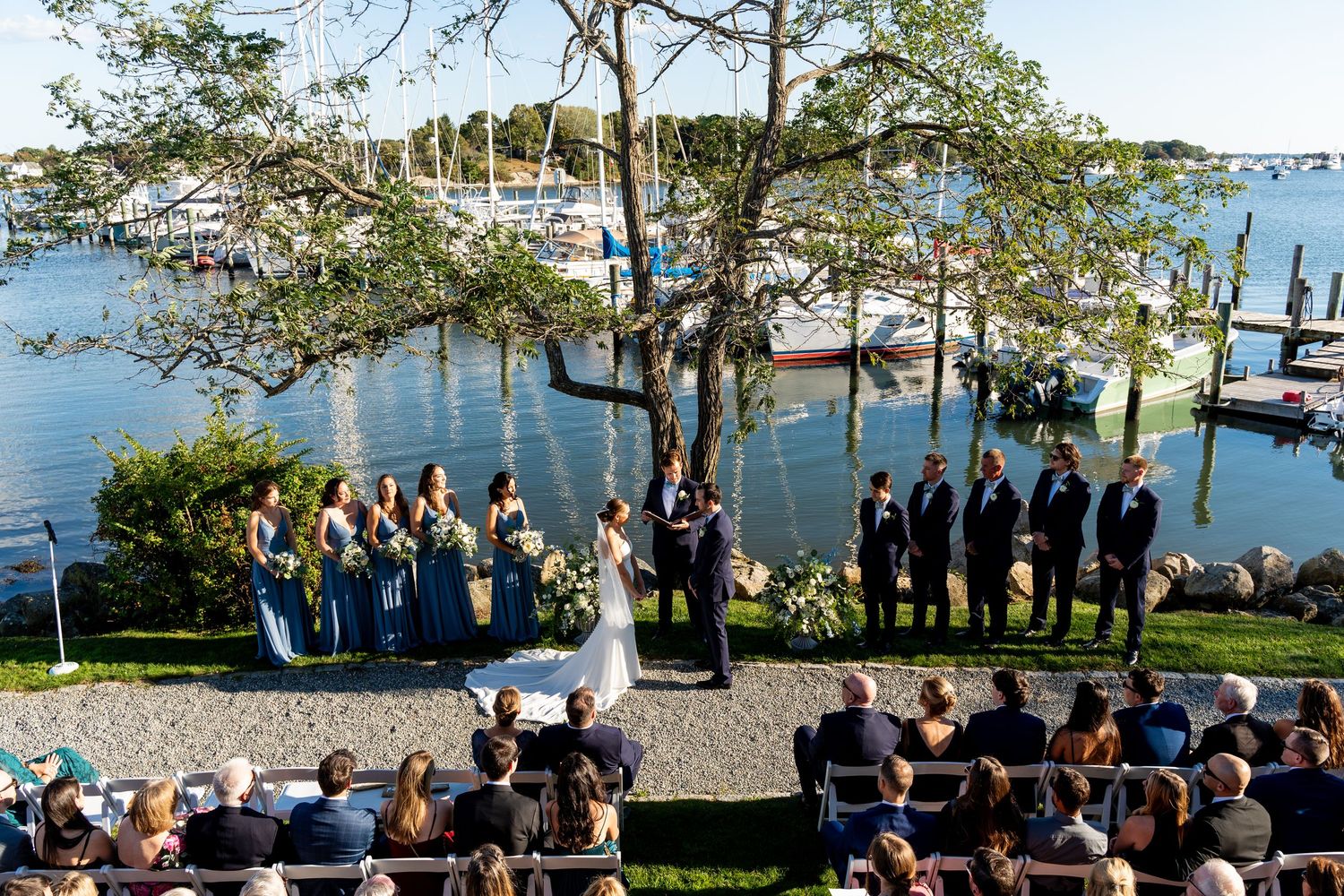 Aerial view of waterfront wedding ceremony with guests seated by marina and boats.