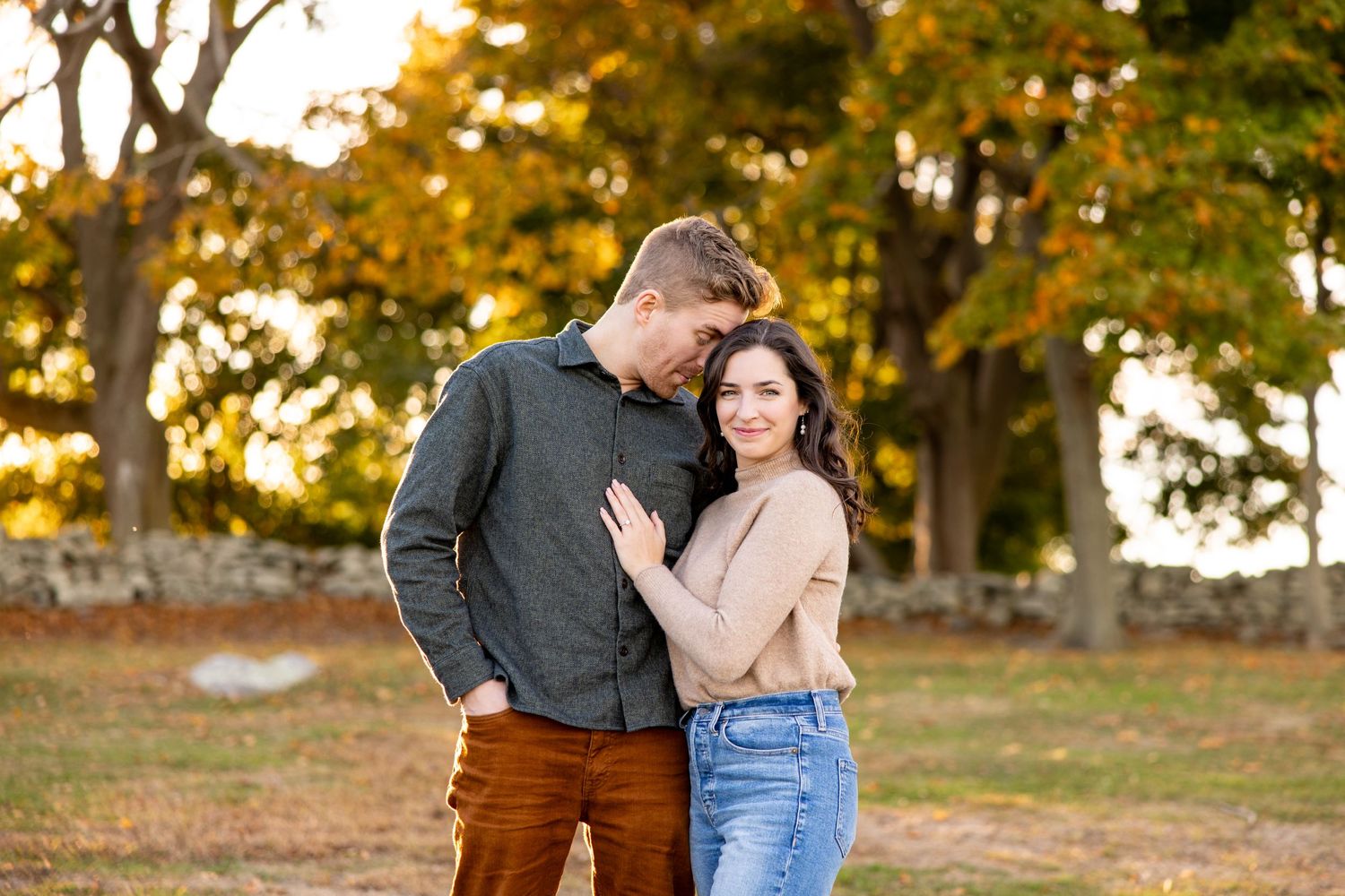 A romantic couple embraces in casual attire against an autumn backdrop with golden trees at sunset.