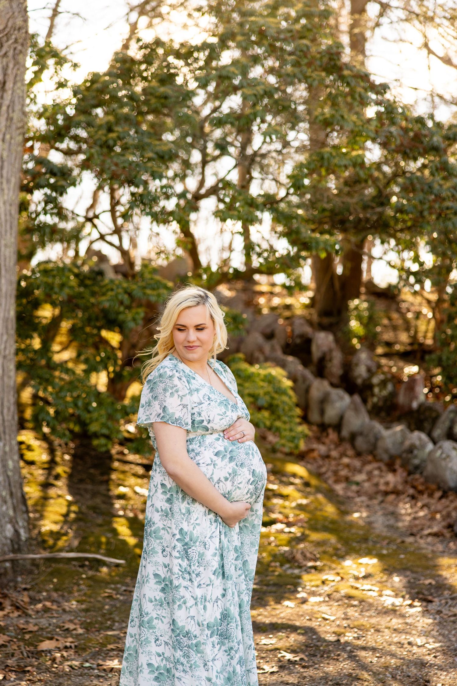 Expectant mother in a floral dress poses in a sunlit wooded setting.