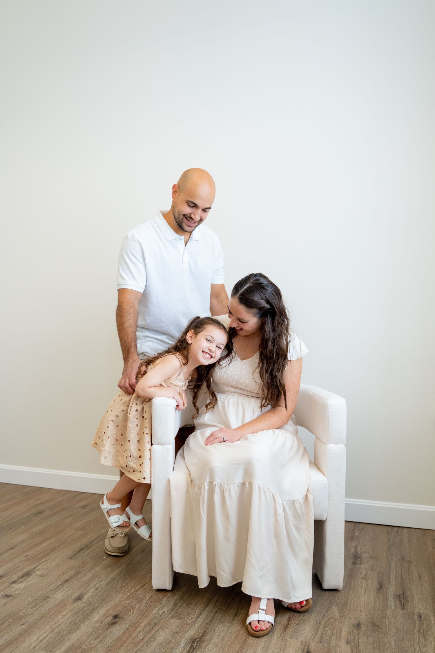 Family of three sharing tender moment while seated in white chair against neutral background.