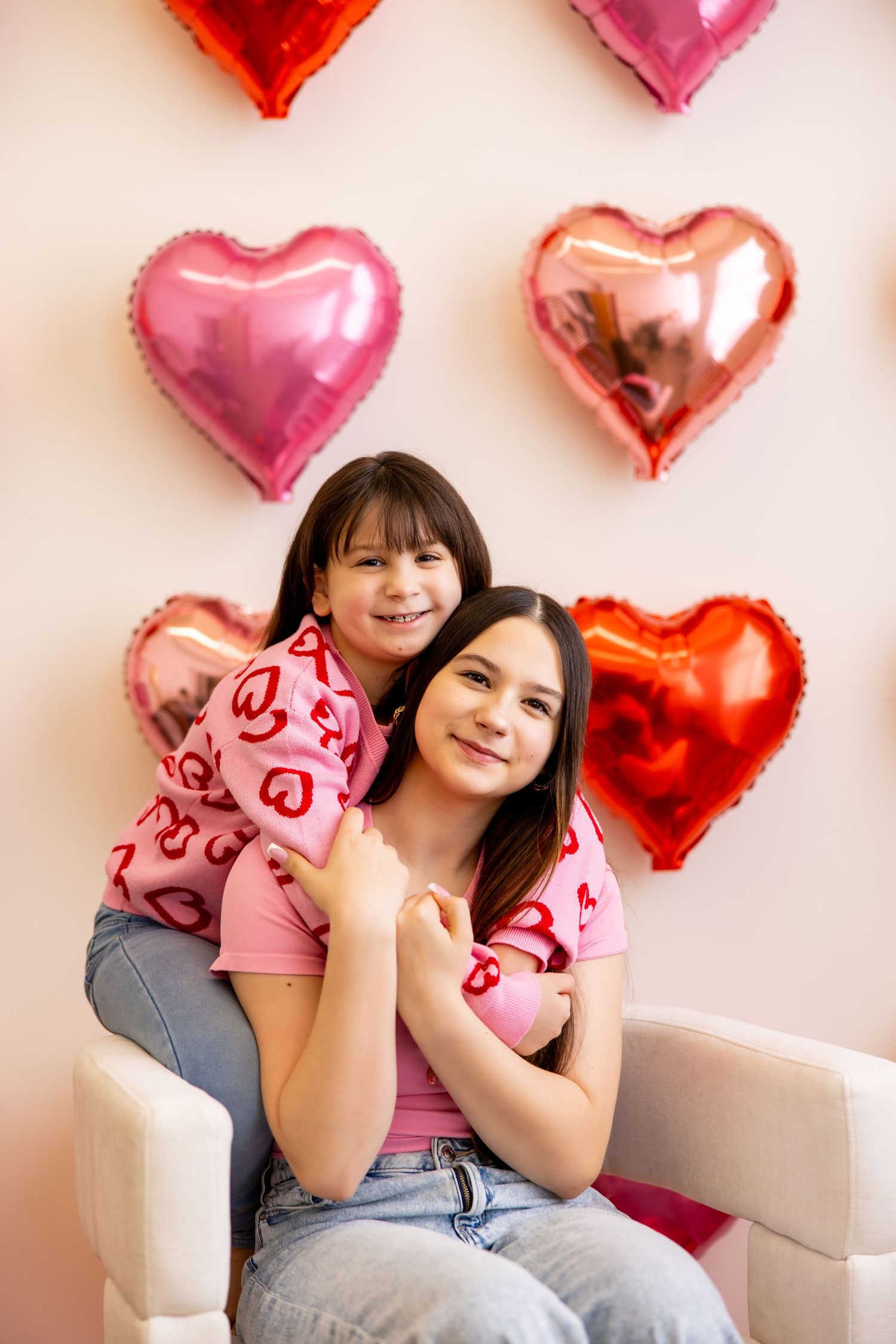 Two people pose on white chair with heart-shaped balloons decorating the background.