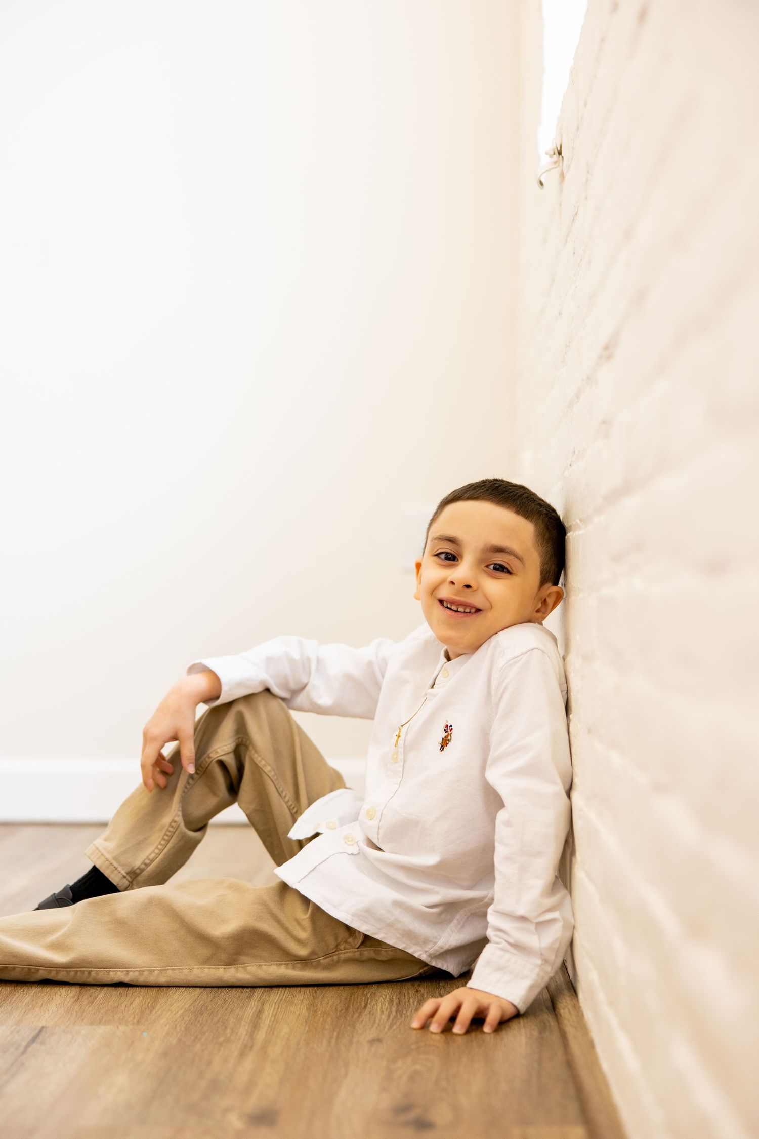 A casual portrait of a young person in white shirt and khakis sitting against white wall.