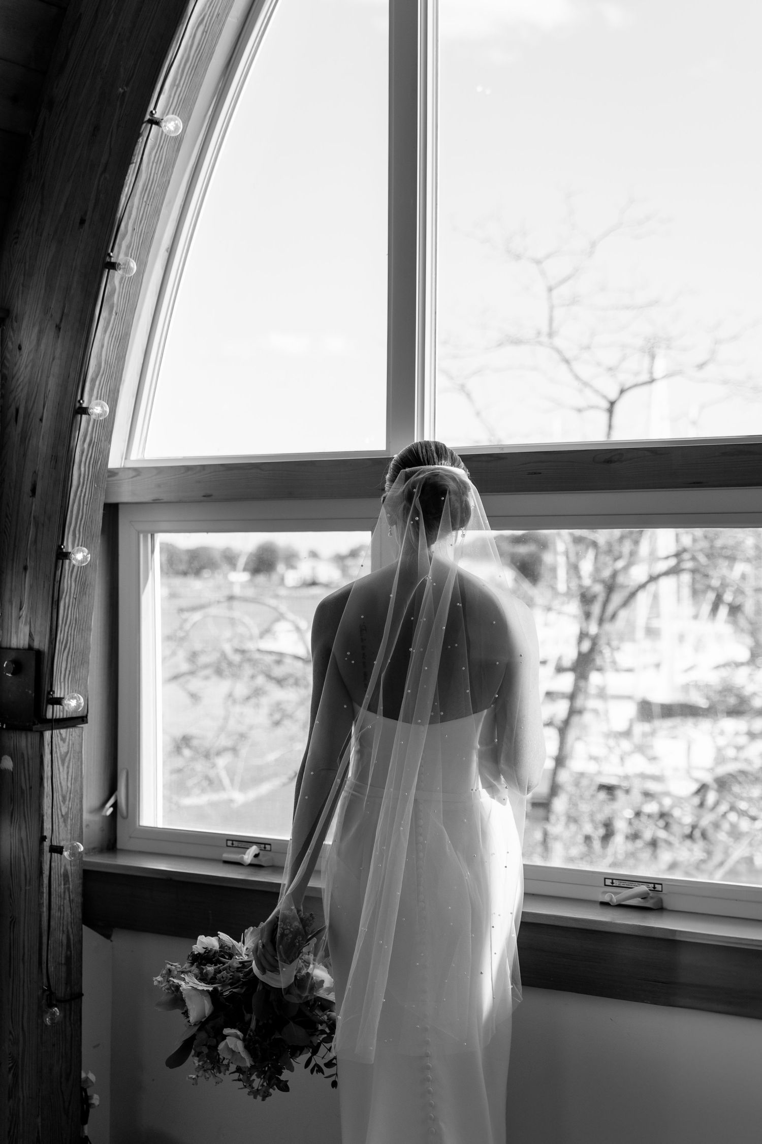 Silhouette of bride in white dress looking through arched window in black and white.