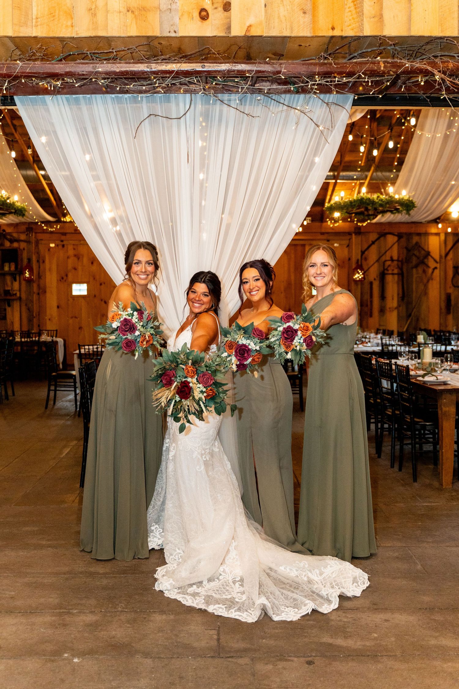 Bride poses with bridesmaids in sage green dresses holding burgundy bouquets in rustic venue.