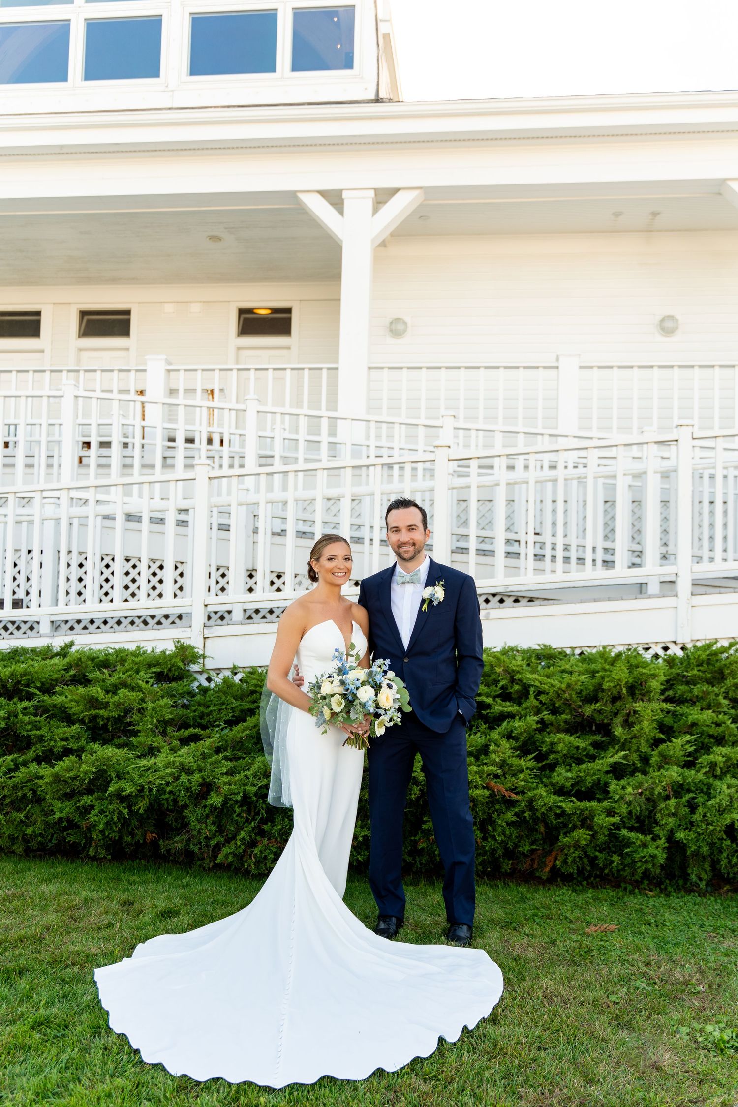 Wedding couple poses in formal attire against white building with classic architectural details.