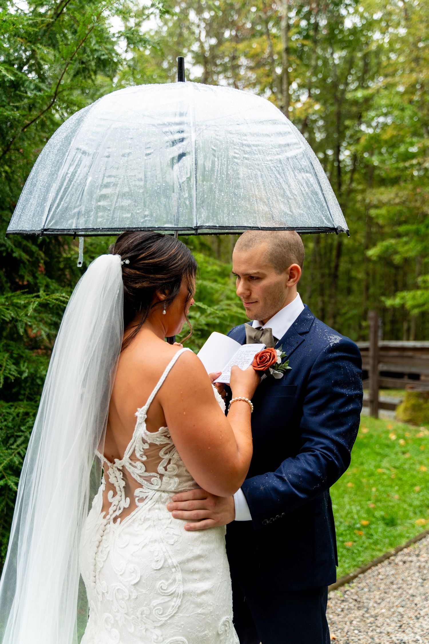 Newlyweds reading vows while standing close under a clear umbrella in a garden.