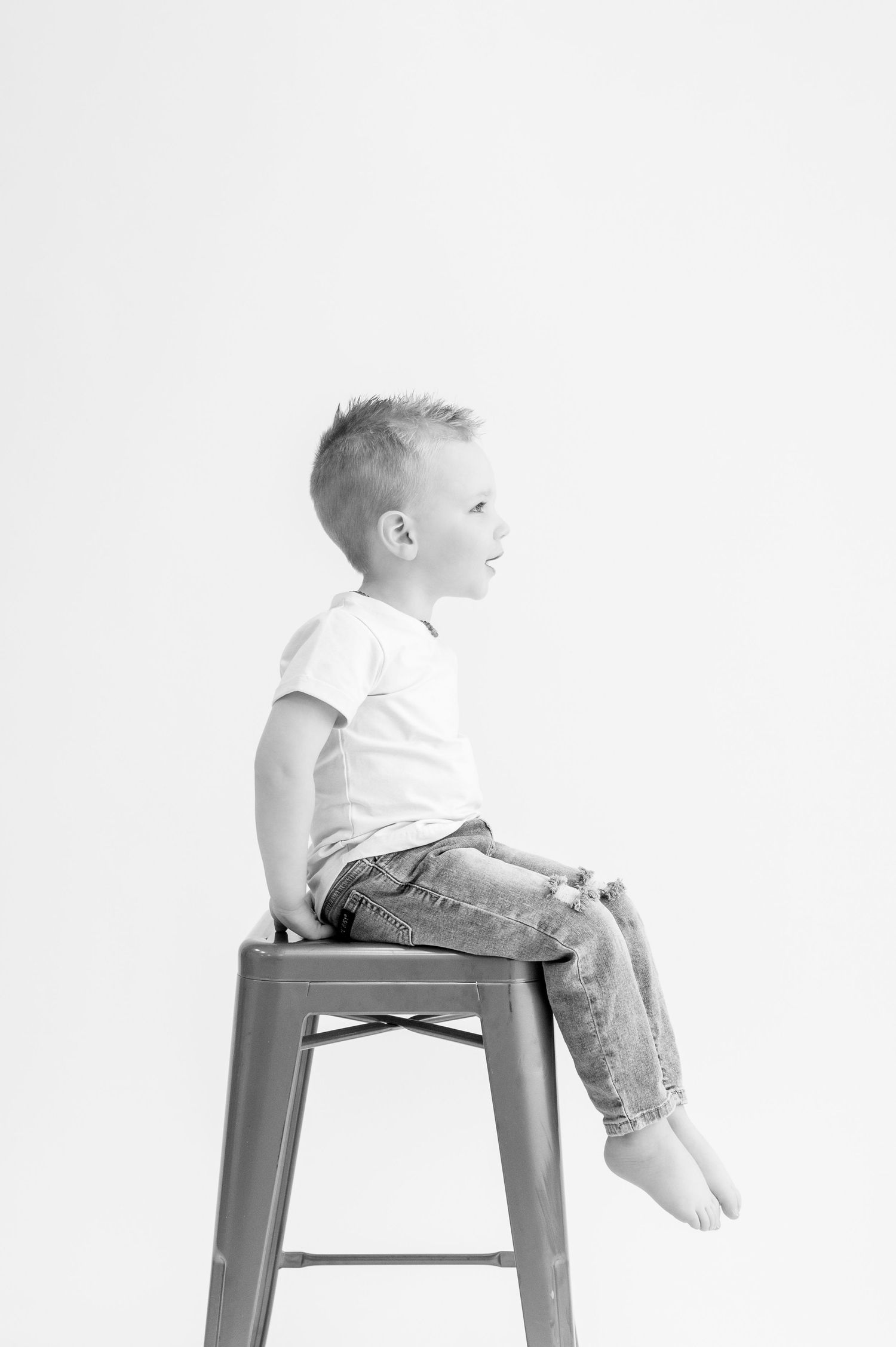Child in white shirt sitting on metal stool against white background in black and white photo.