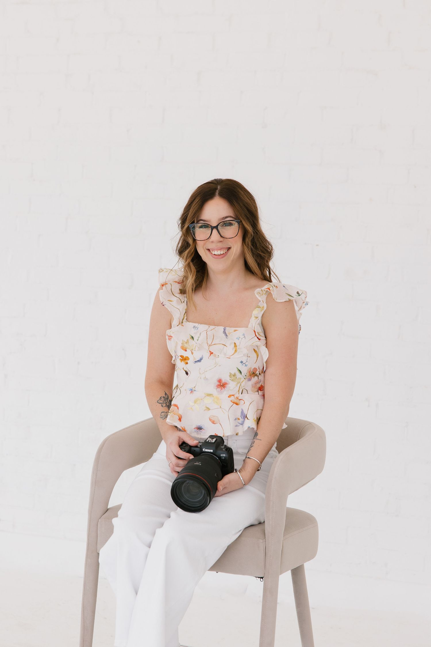 Professional photographer wearing glasses and floral top sits in light gray chair holding camera against white backdrop.