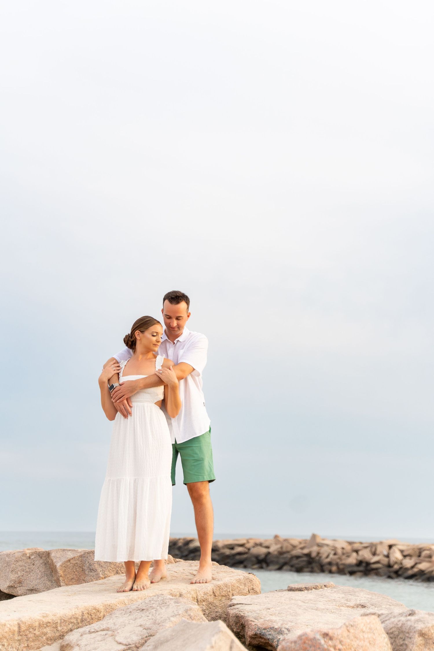 A couple stands together on rocky coastal outcropping against bright sky.