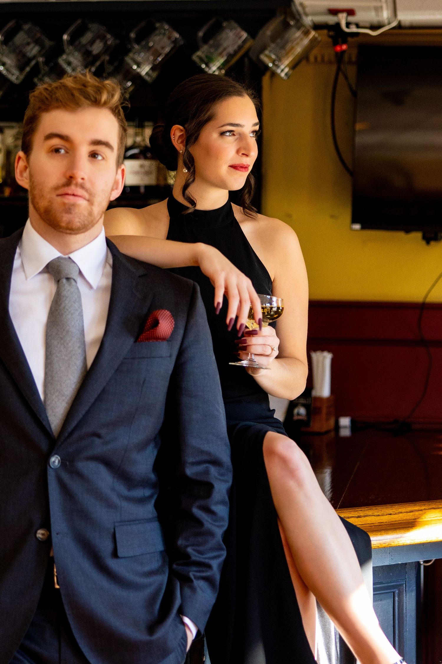 Elegant couple poses at a bar, with a woman in black dress and man in navy suit with red pocket square.