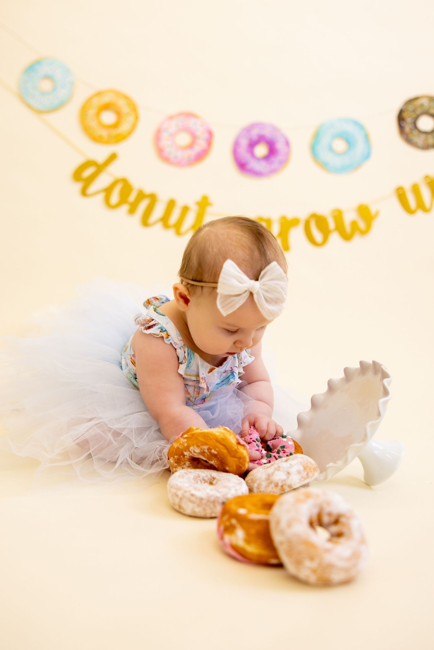 Baby in tulle dress reaches for donuts under colorful donut-themed banner.