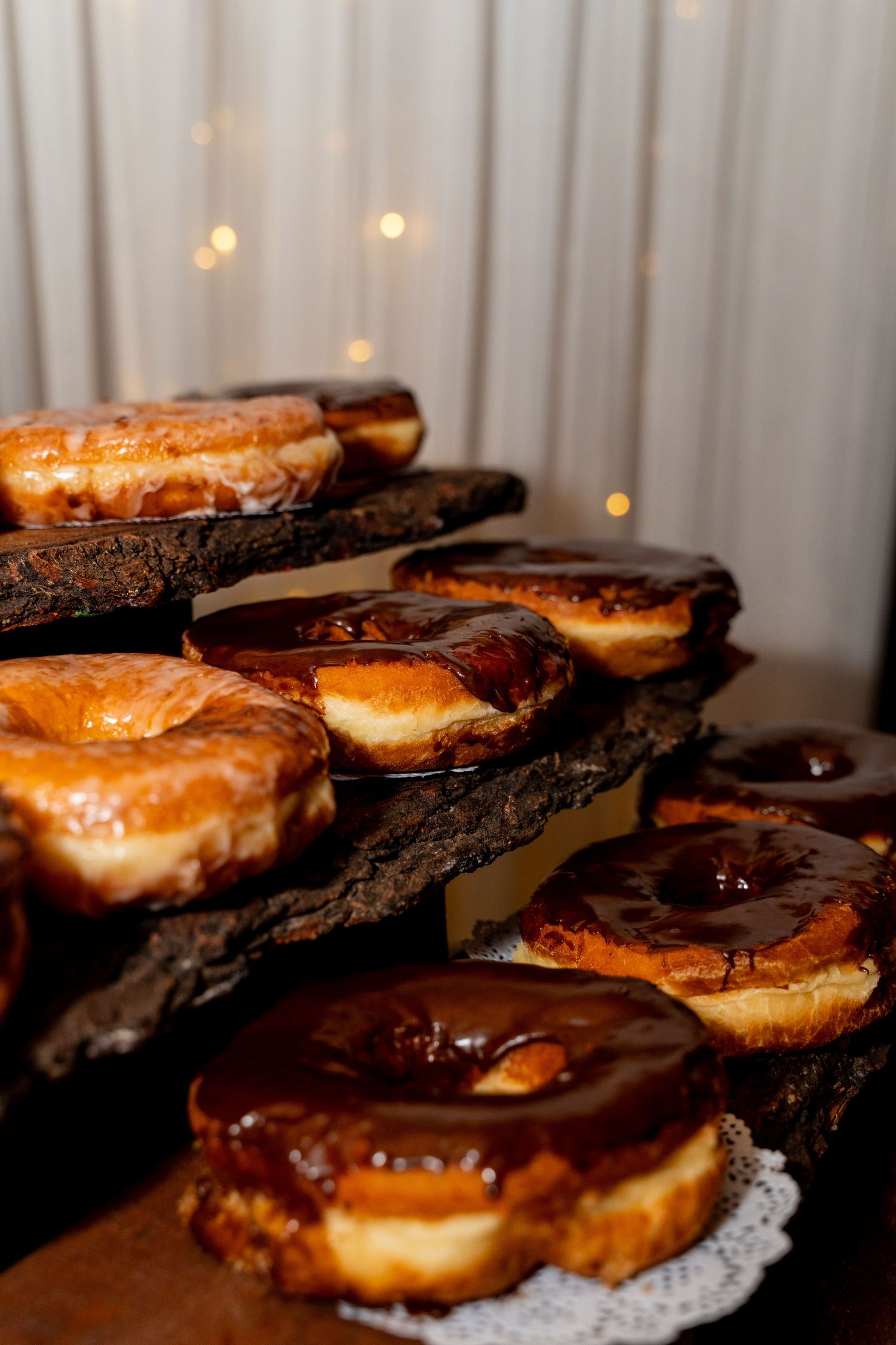 Chocolate glazed donuts displayed on rustic wooden tiers with twinkling lights in background.