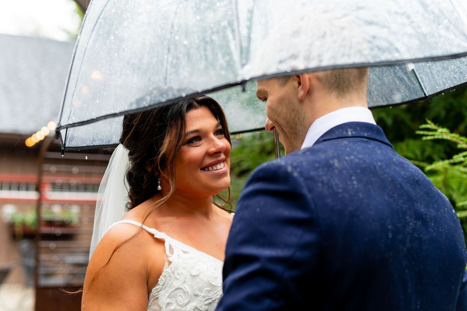 Close-up shot of newlyweds sharing a joyful moment under a clear umbrella against greenery.