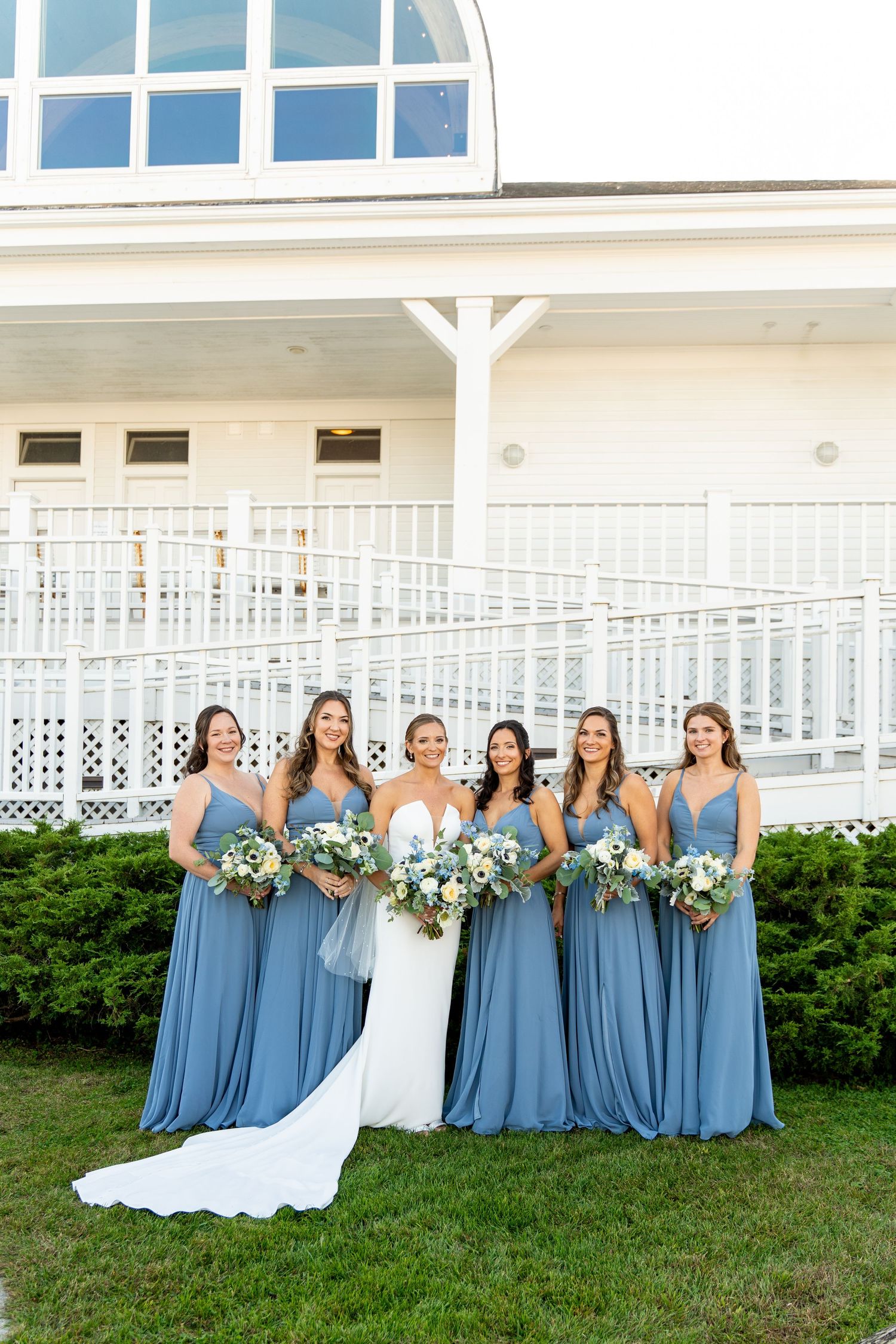 Bridesmaids in dusty blue dresses pose with bouquets beside bride in white gown against white building exterior.