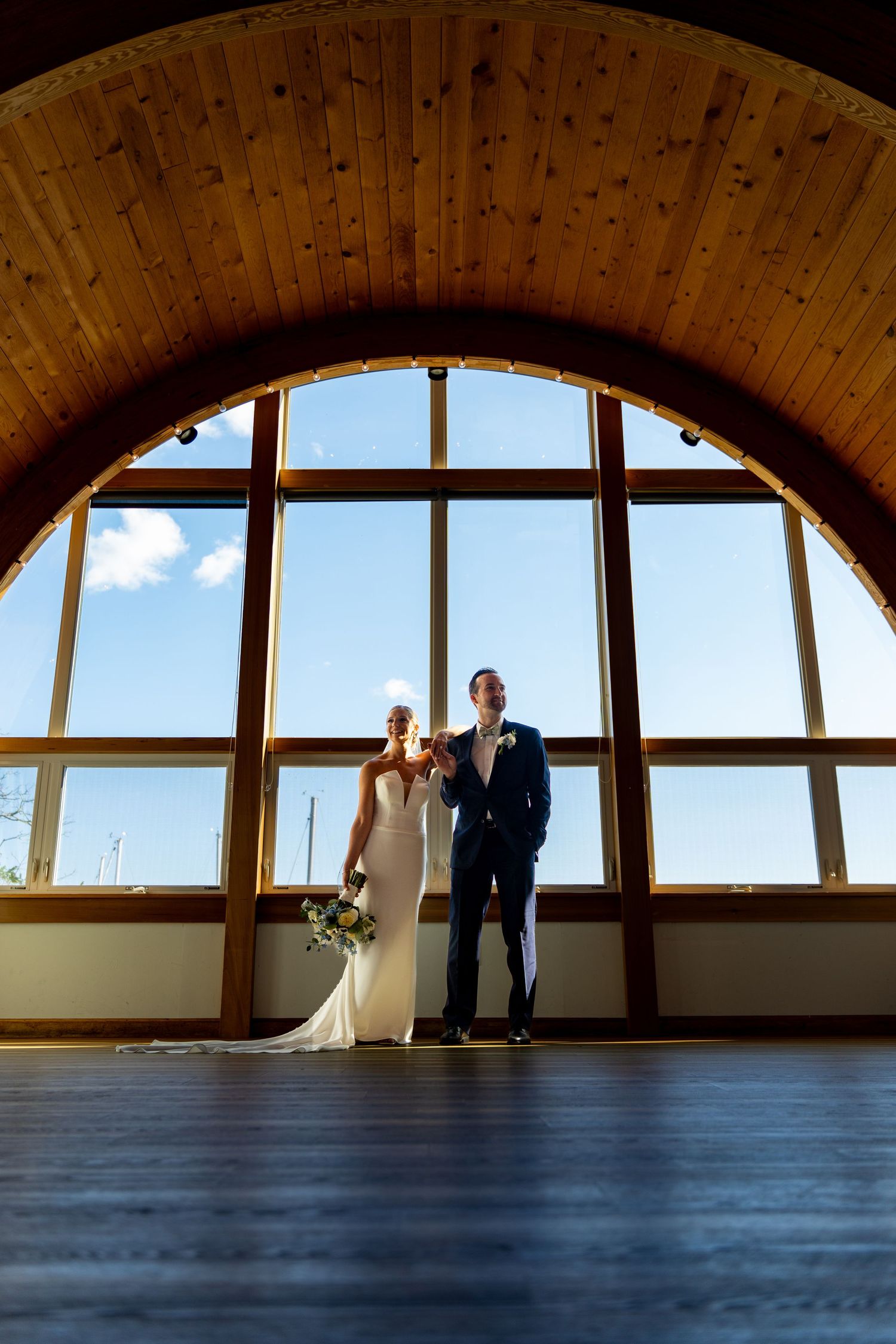 Wedding couple silhouetted against large arched window with wooden ceiling beams.