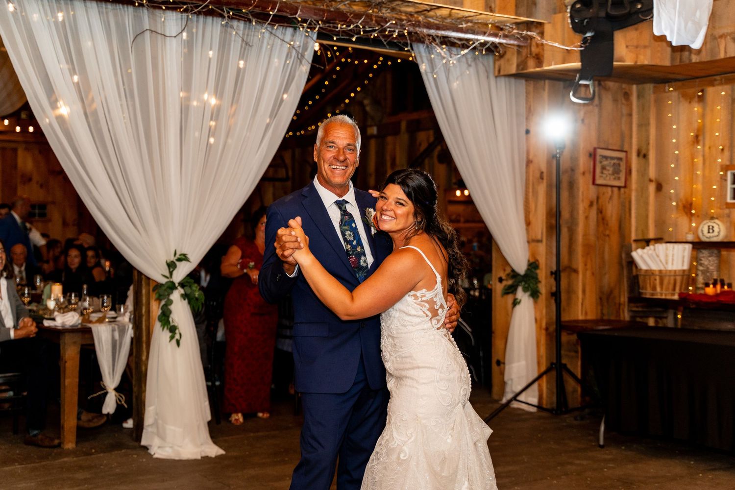 A couple dances in a rustic barn venue decorated with white drapes and twinkling lights.