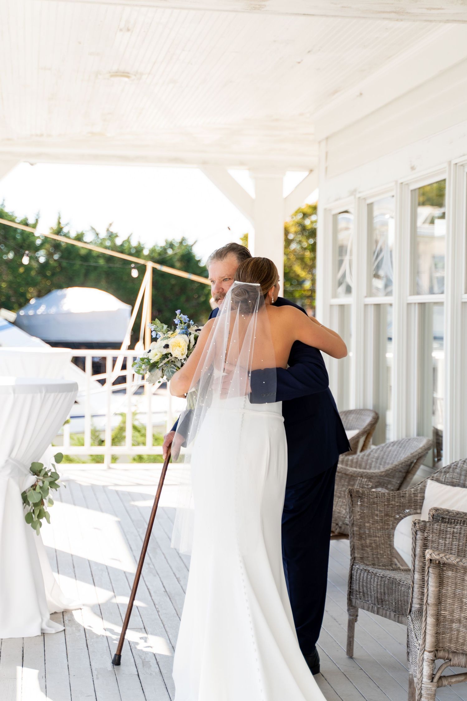 Two people embrace on a white porch with wicker furniture and a flowing white veil.