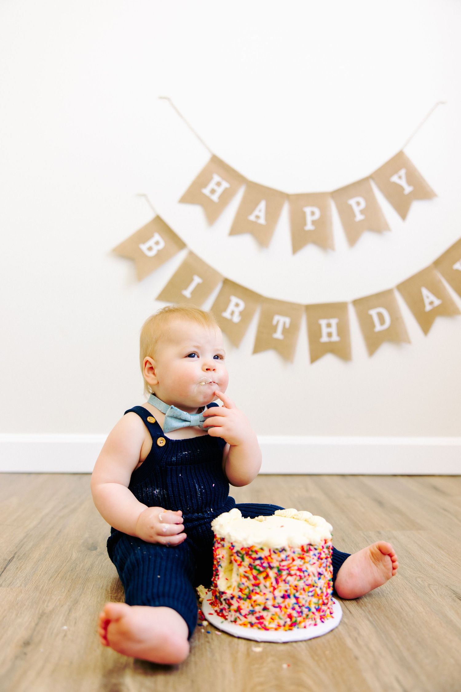 Baby in blue overalls sitting beside festive birthday cake with burlap banner decoration.